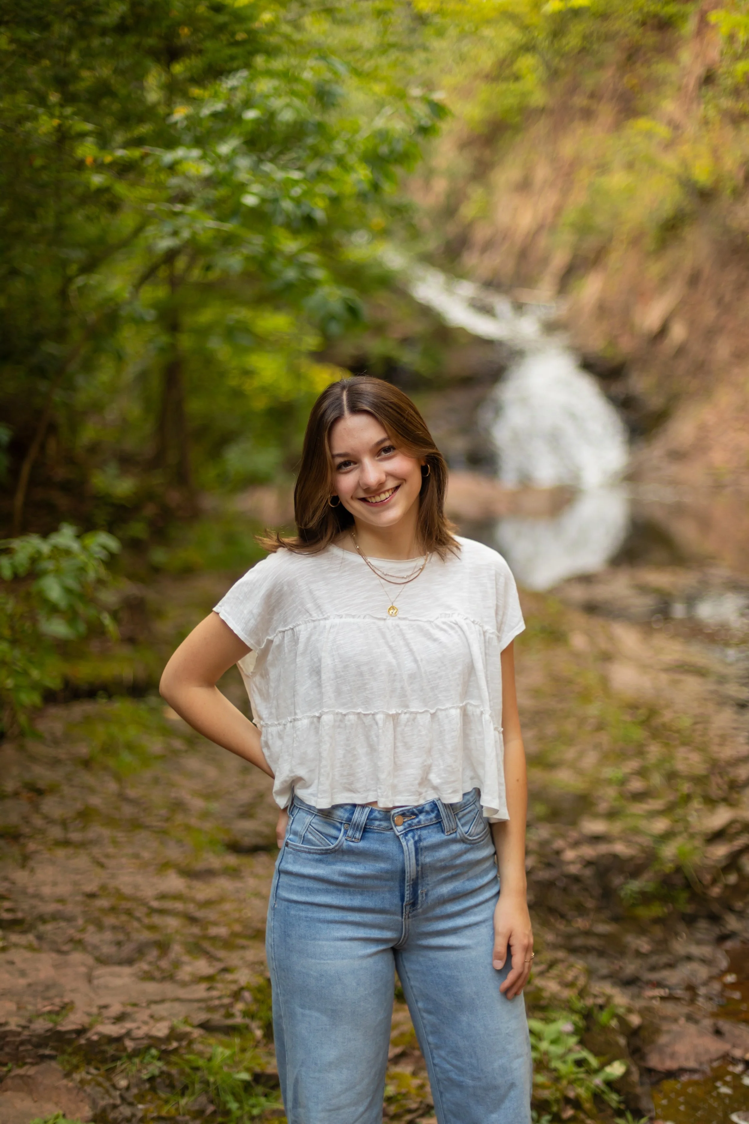 A young woman standing outdoors in front of a waterfall and surrounded by greenery, smiling at the camera.