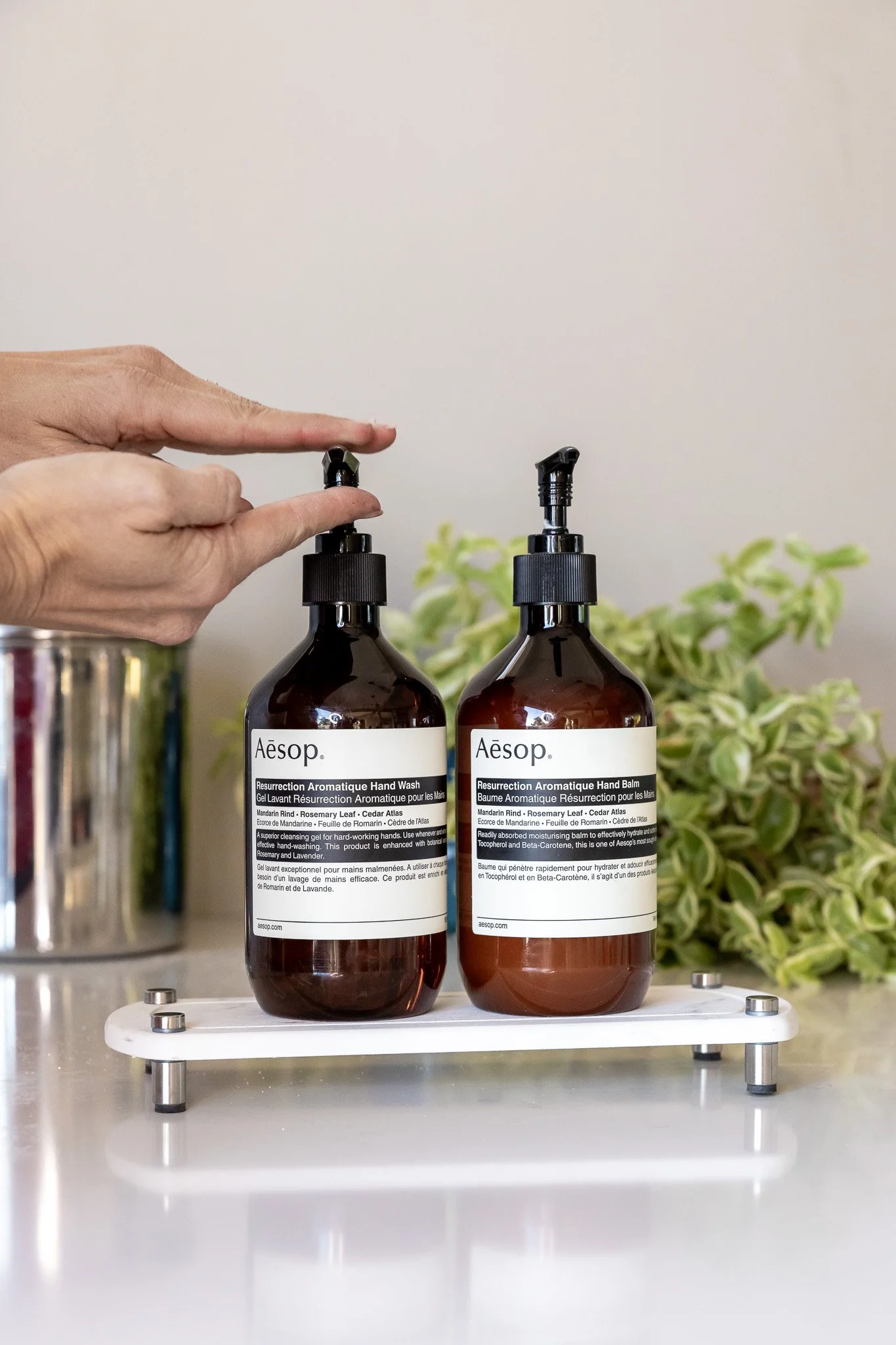 Person's hand pressing top of a dark amber dispenser bottle labeled 'Aesop Resurrection Aromatique Hand Wash' on a white tray, with another similar bottle labeled 'Aesop Resurrection Aromatique Hand Balm' beside it, and a green leafy plant in the bac