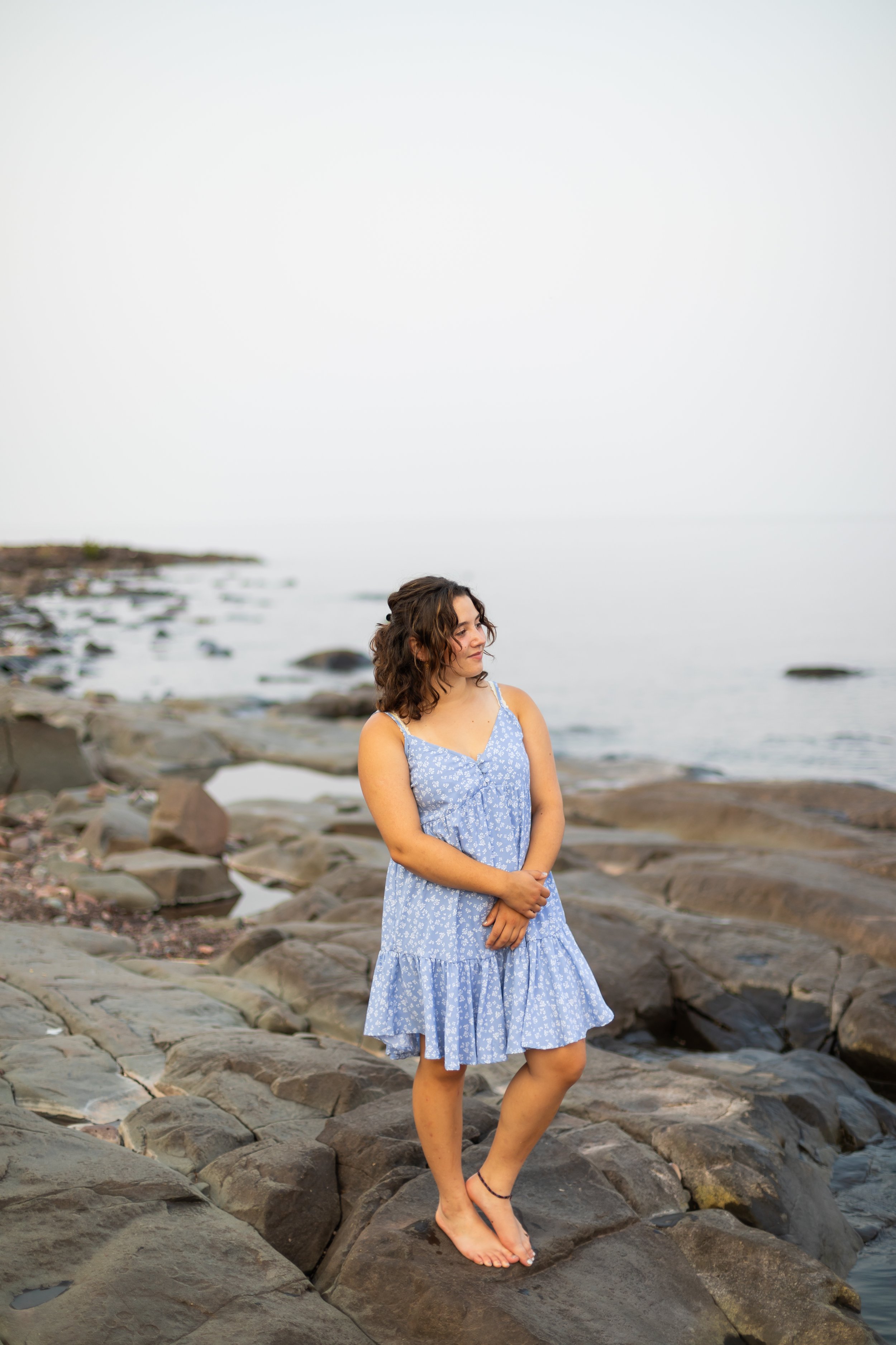 Young woman in a blue floral dress standing barefoot on rocks by the water, looking to the side near a calm shoreline.