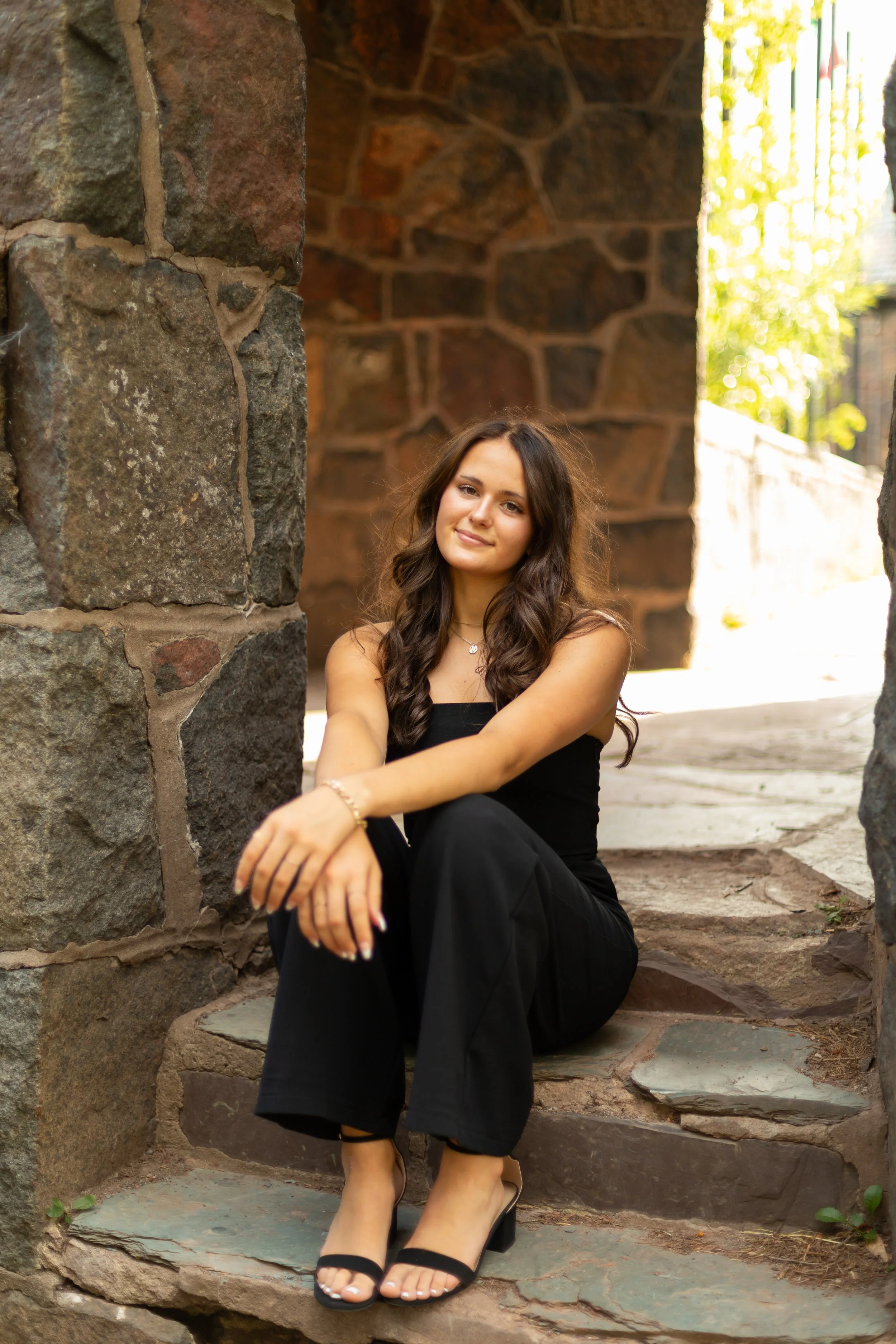 A young woman with long, wavy brown hair, wearing a black sleeveless top, black pants, and black high-heeled sandals, sitting on stone steps between two stone walls outdoors, smiling gently, with sunlight filtering through trees.