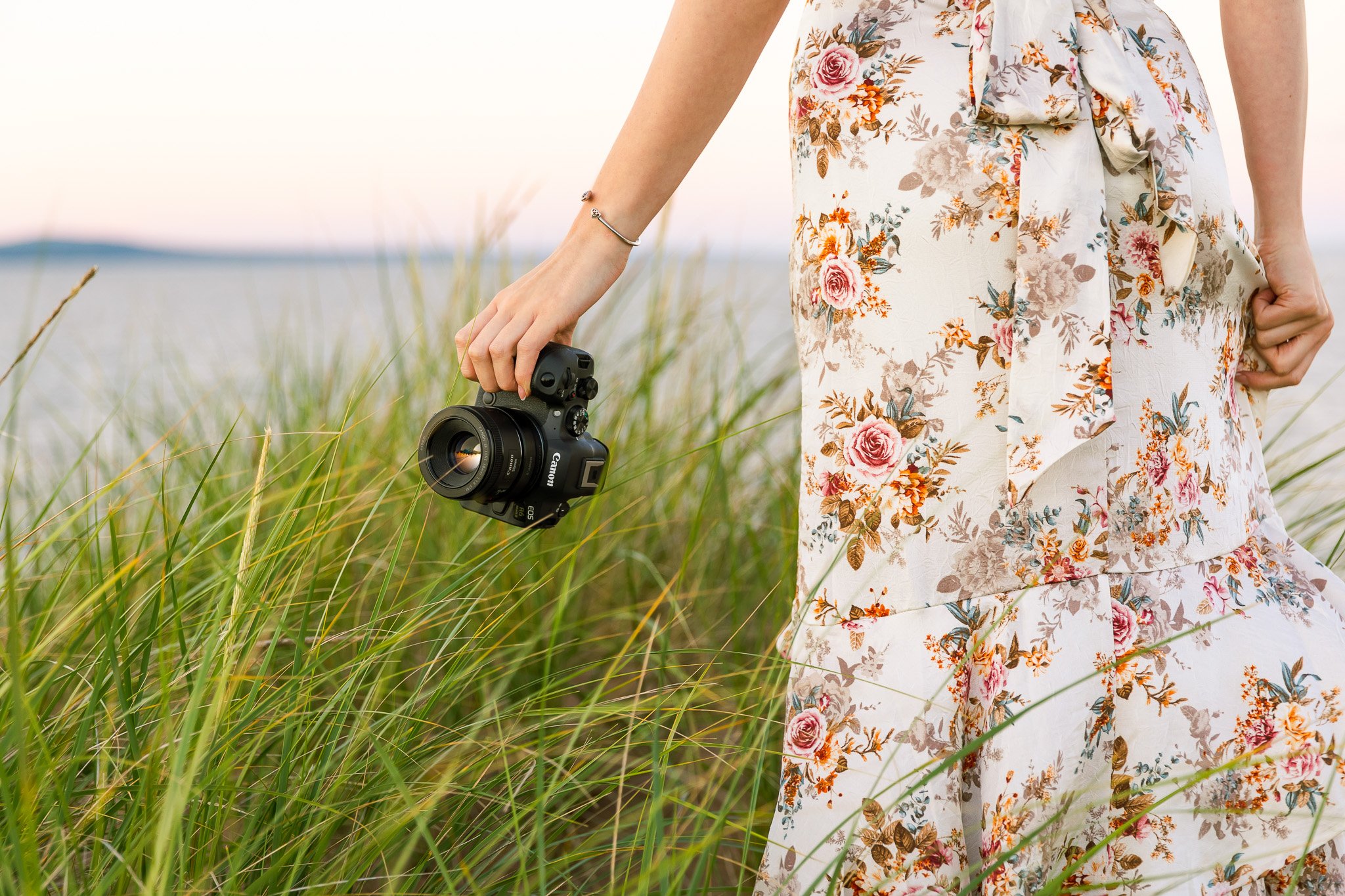 A woman in a floral dress holding a Canon camera while standing in a grassy field near water during sunset.