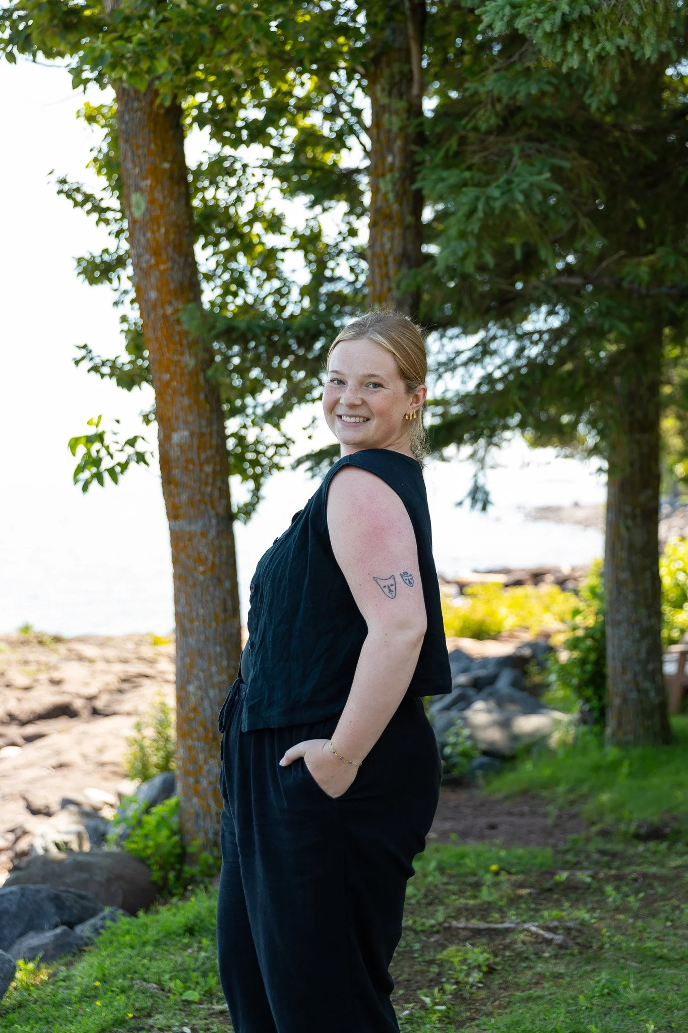 Young woman with blonde hair smiling outdoors near trees and a water body, wearing a black sleeveless top, with tattoos on her arm, and her hands in her pockets.