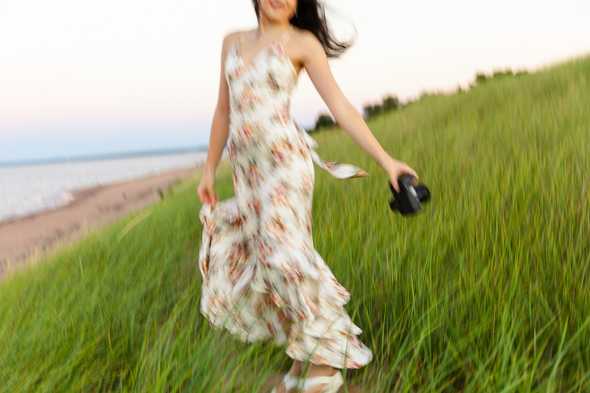A woman in a floral dress is walking through a grassy field near the beach, holding a camera in her right hand with her hair flowing in the breeze during sunset.