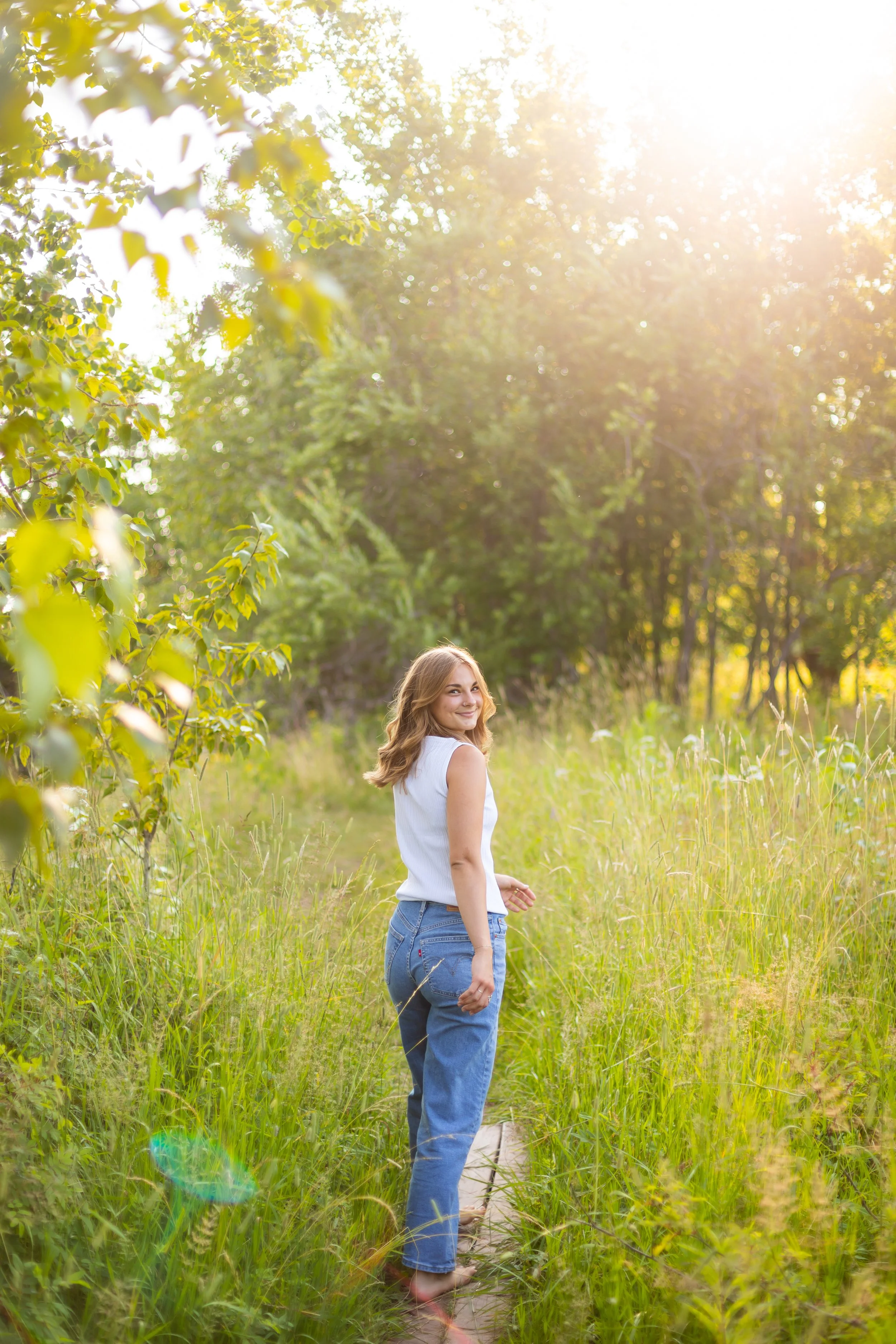 A woman walking barefoot on a narrow path through tall grass in a sunlit forest or park, smiling at the camera.