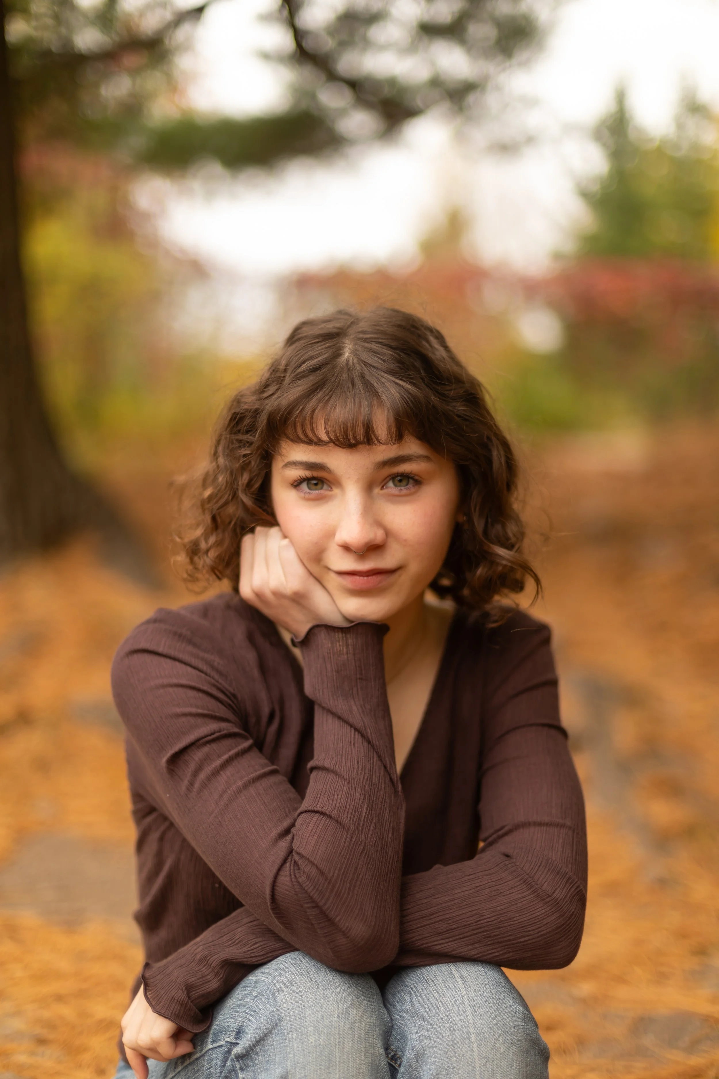 A young woman with curly brown hair and fair skin sitting outdoors on a fall day, resting her chin on her hand, with autumn leaves and trees in the background.