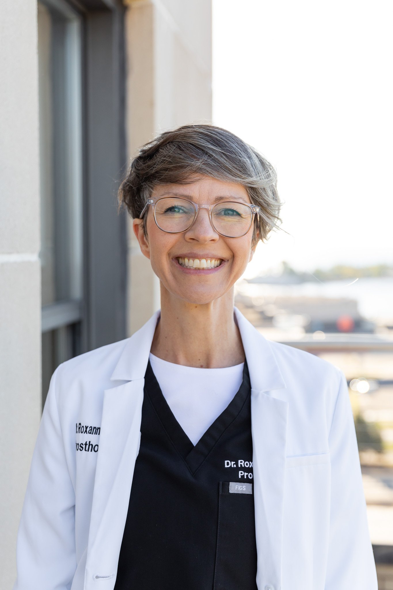Portrait of a smiling woman with short gray hair, glasses, wearing a white medical coat and black scrubs, standing outdoors near a building with a blurred city background.
