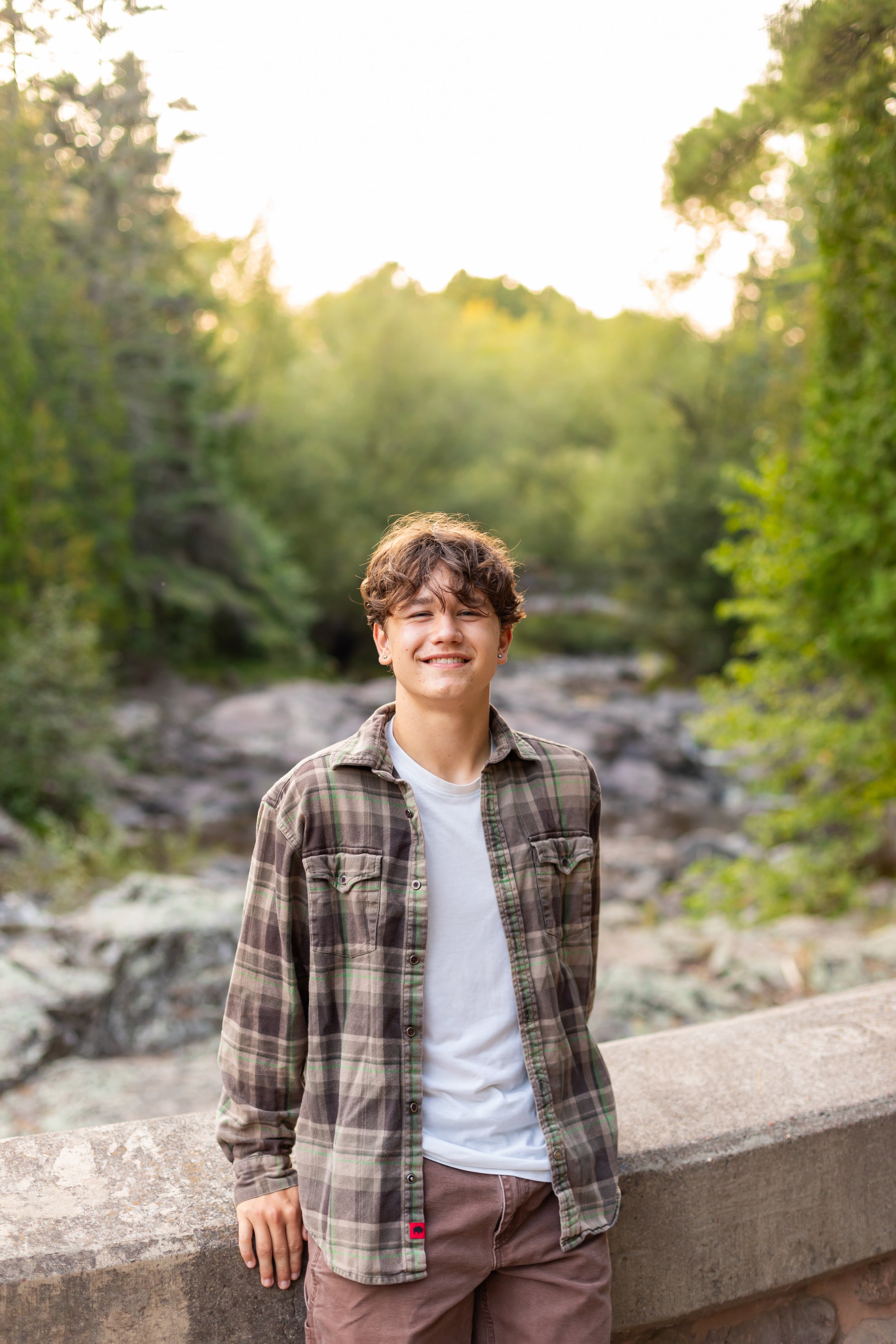 A smiling young man with curly hair, wearing a plaid shirt over a white t-shirt, standing outdoors by a stone railing with a forested area and creek in the background during golden hour.