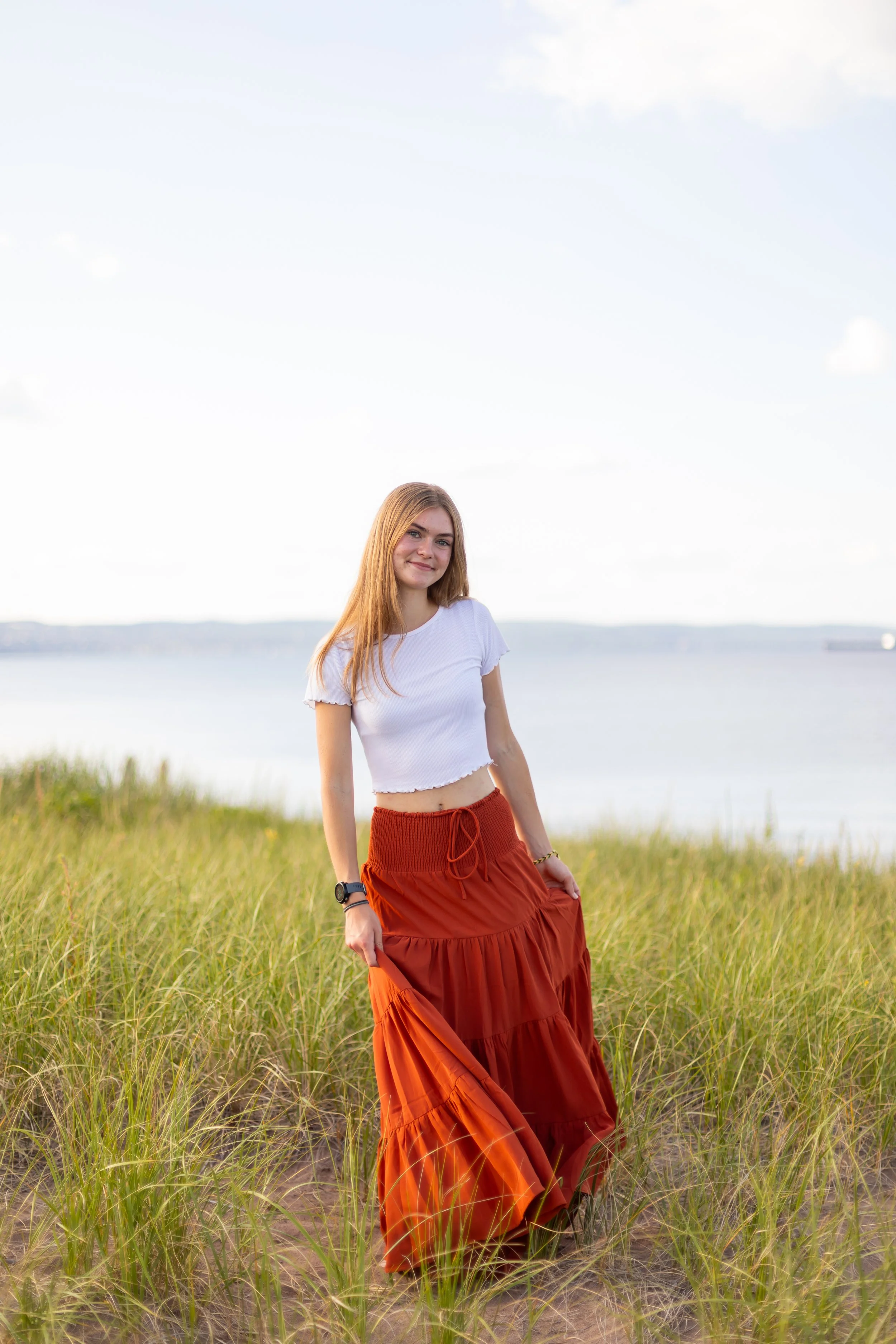 A young woman with red hair stands in a grassy field near a body of water on a clear day, wearing a white crop top and a long orange skirt.