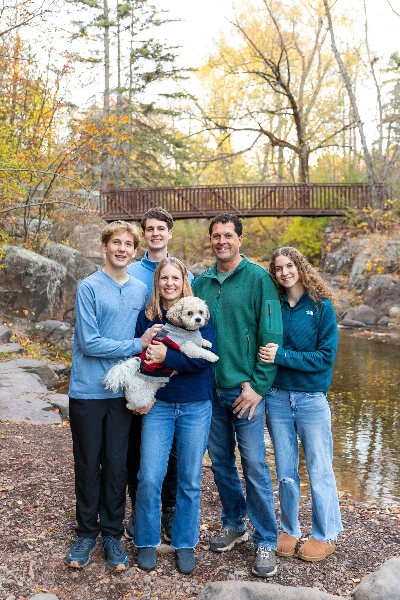 A family of five, including a dog, standing outdoors near a creek with autumn trees and a wooden bridge in the background.