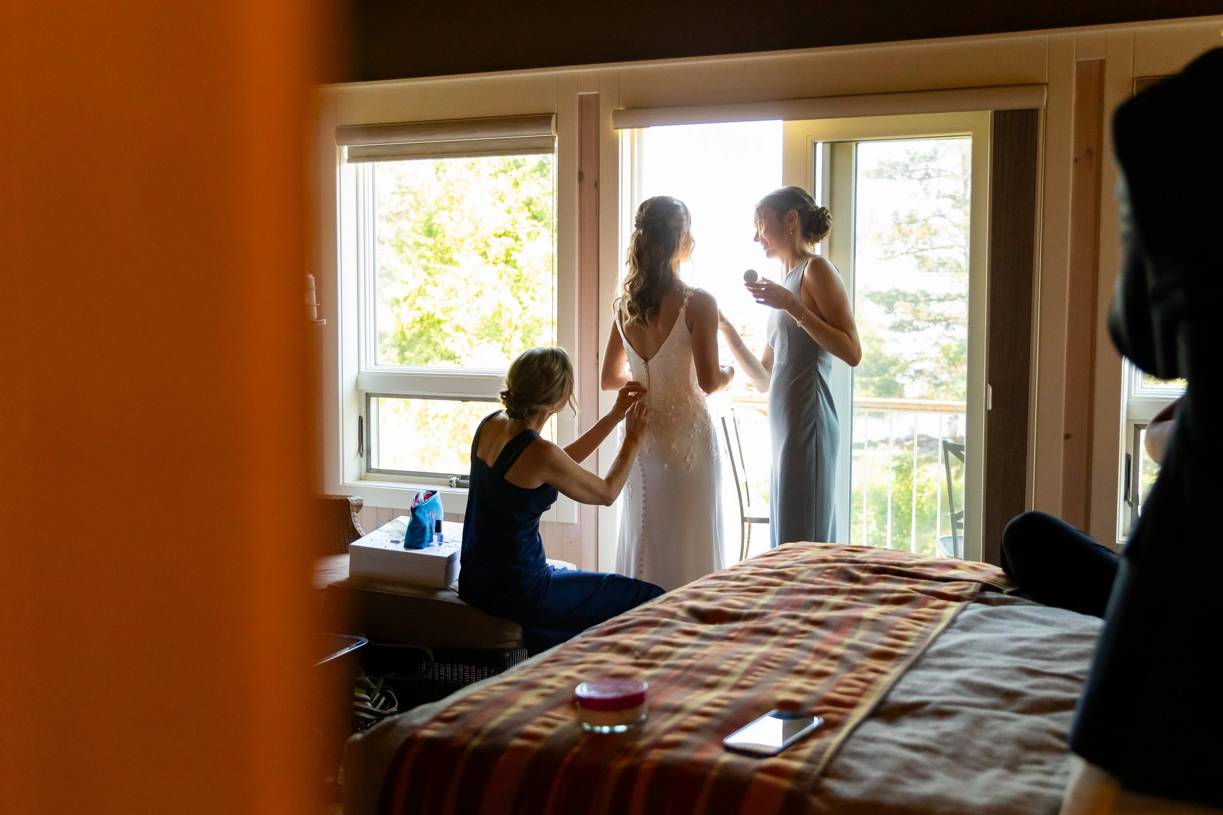Women preparing a bride for her wedding in a bright bedroom with large windows, a woman in a dark dress helping with the dress, and another woman in a light-colored dress holding a makeup brush.
