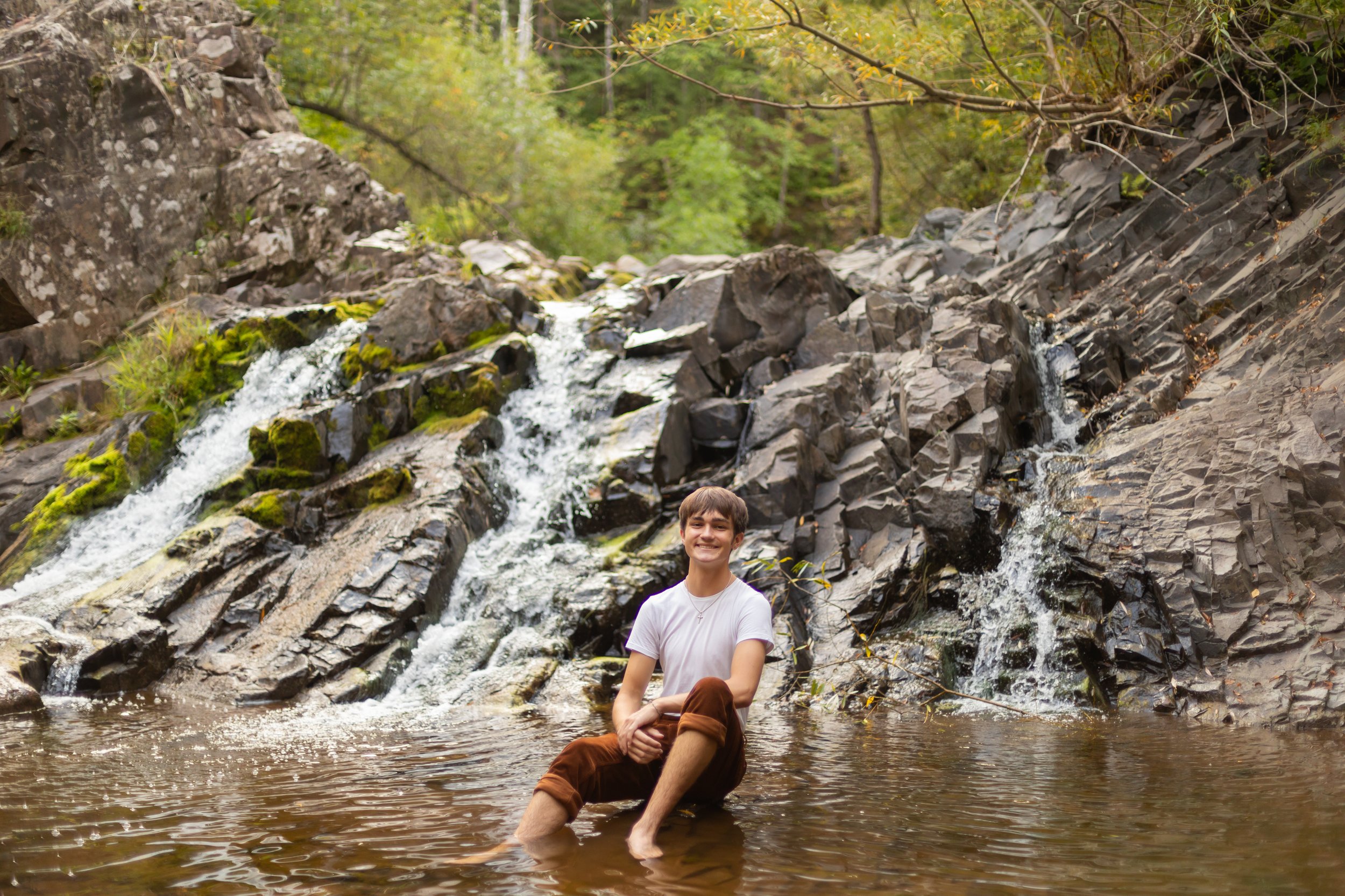 A young man sitting in a shallow creek with small waterfalls, surrounded by green trees and rocks.