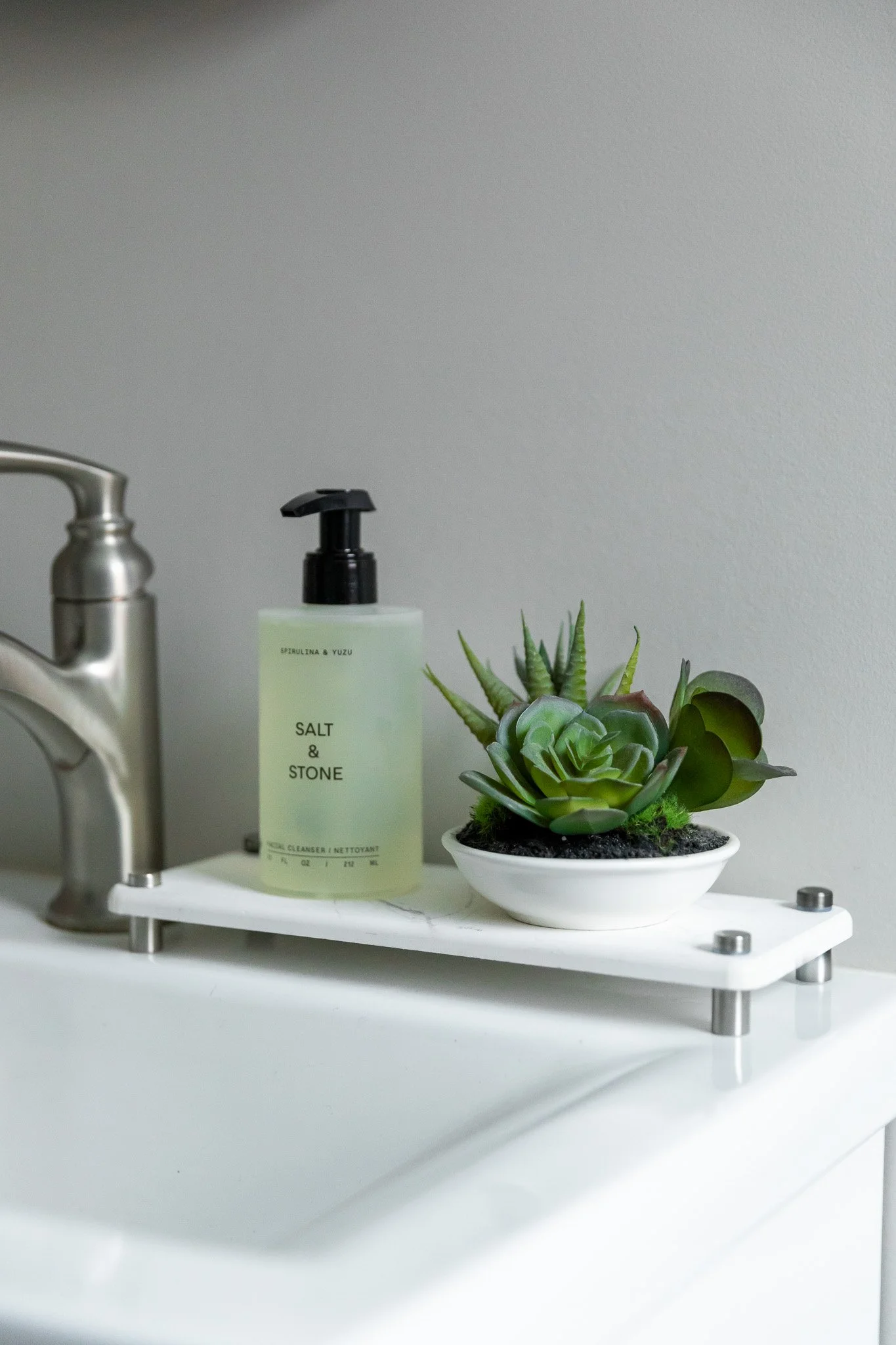 A white shelf with a soap dispenser labeled 'Salt & Stone' and a small green succulent plant in a white bowl, placed on a white kitchen countertop next to a stainless steel faucet.
