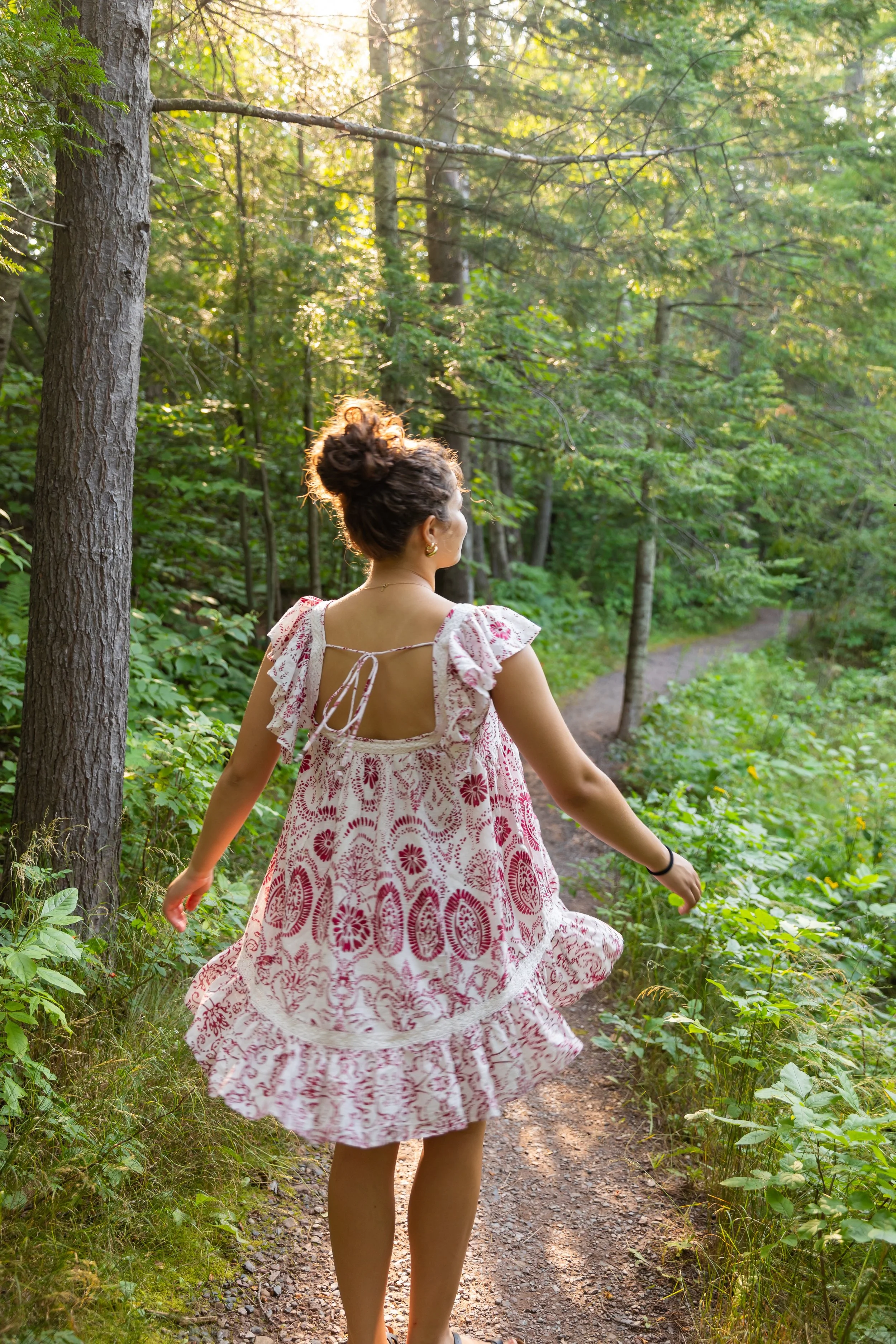 A woman in a pink and white patterned dress walking on a forest trail surrounded by green trees and foliage.