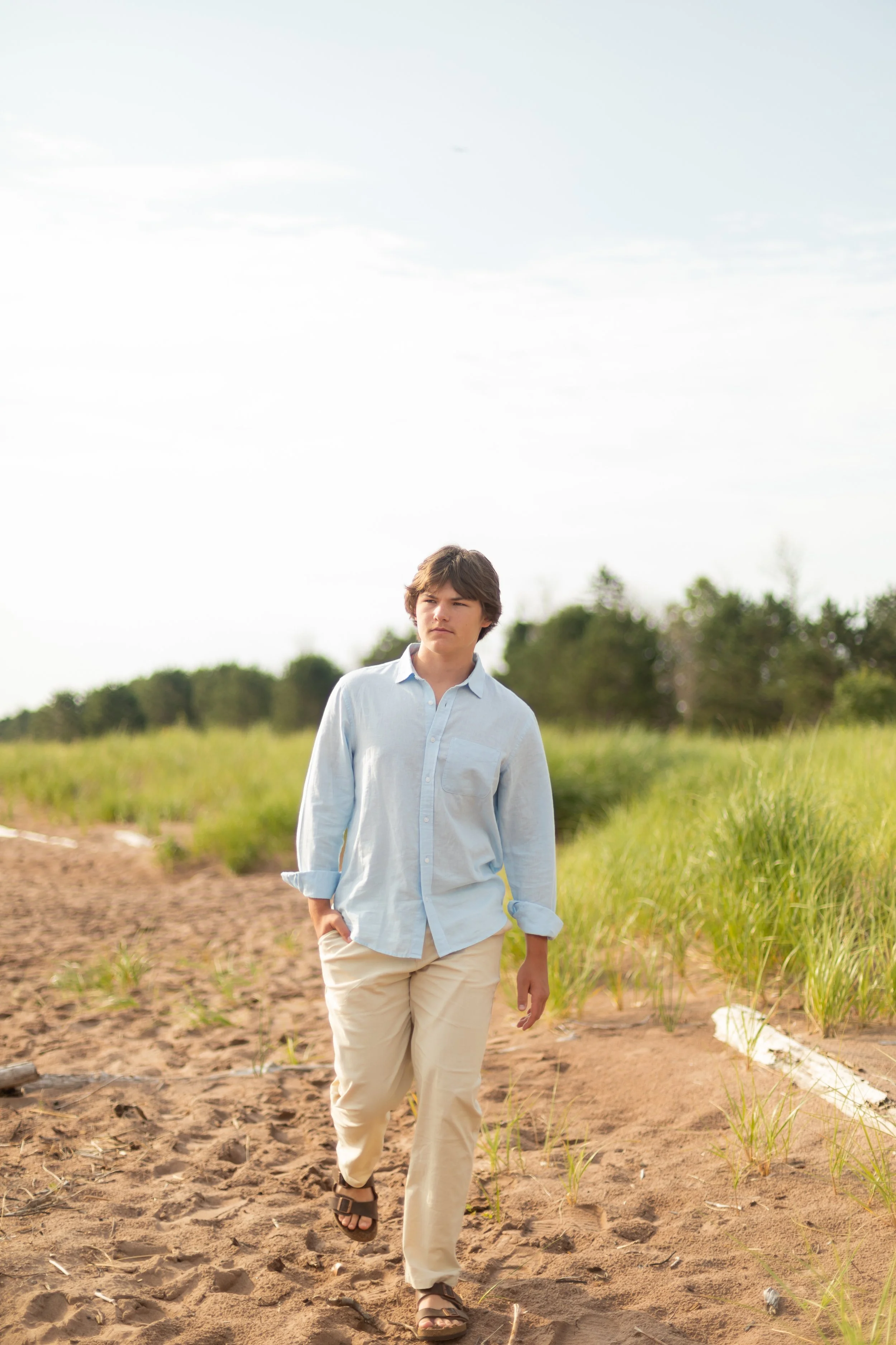 A young man walking on a sandy path through a grassy area on a sunny day, wearing a light blue button-up shirt, beige pants, and sandals.