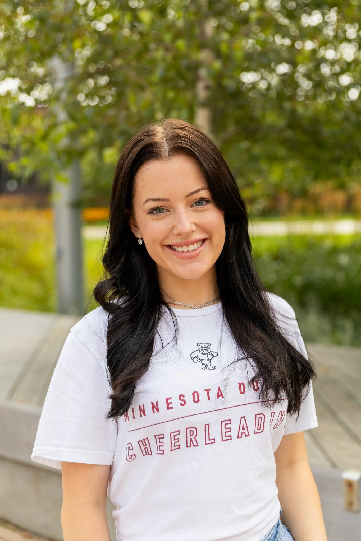 A young woman with long black hair, smiling, wearing a white T-shirt that reads 'Minnesota Duluth Cheerleading' and sitting outdoors with trees and greenery in the background.