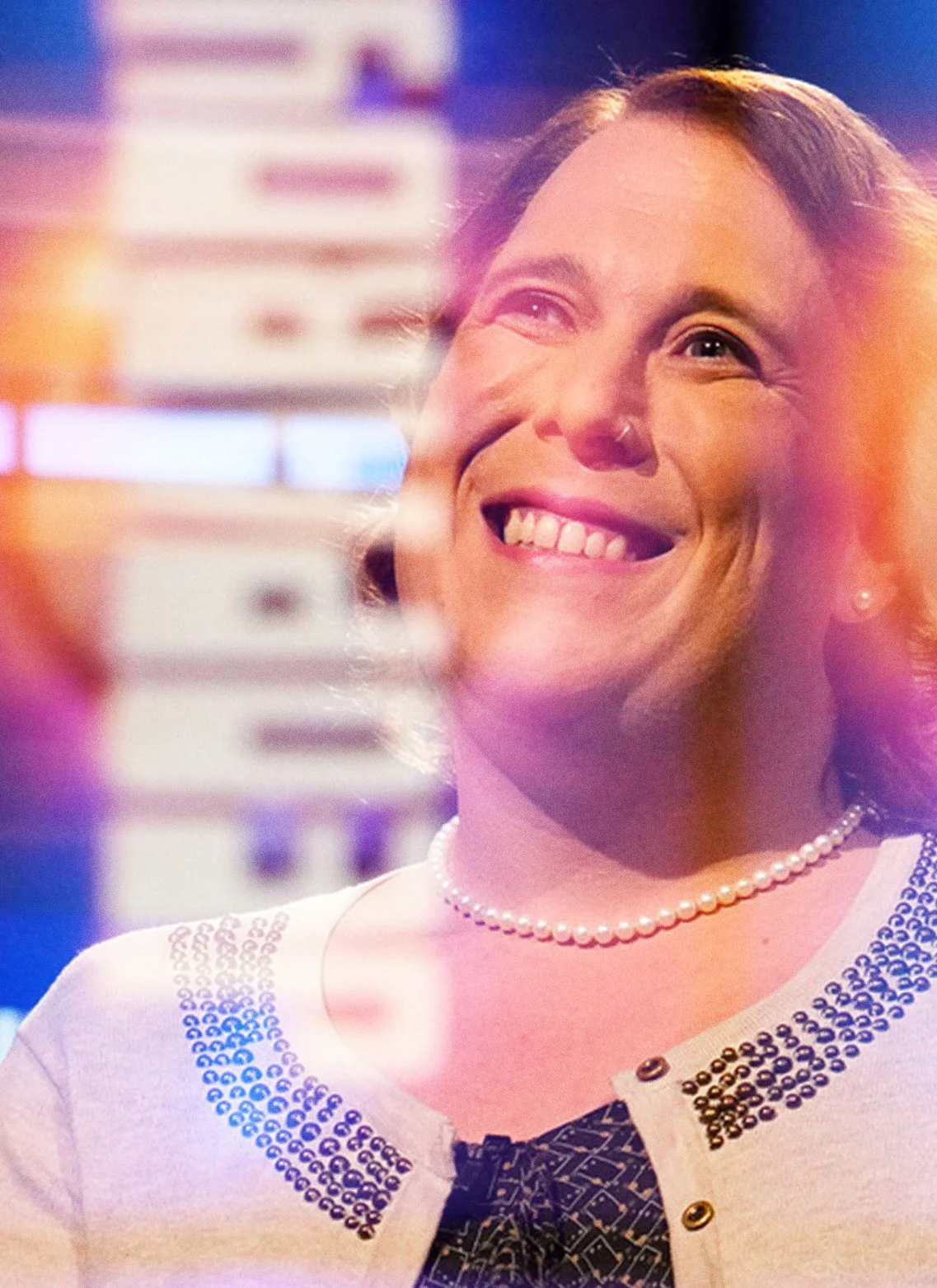 A woman smiling in a brightly lit setting, wearing a pearl necklace and a white top with decorative embellishments, with colorful bokeh lights in the background.