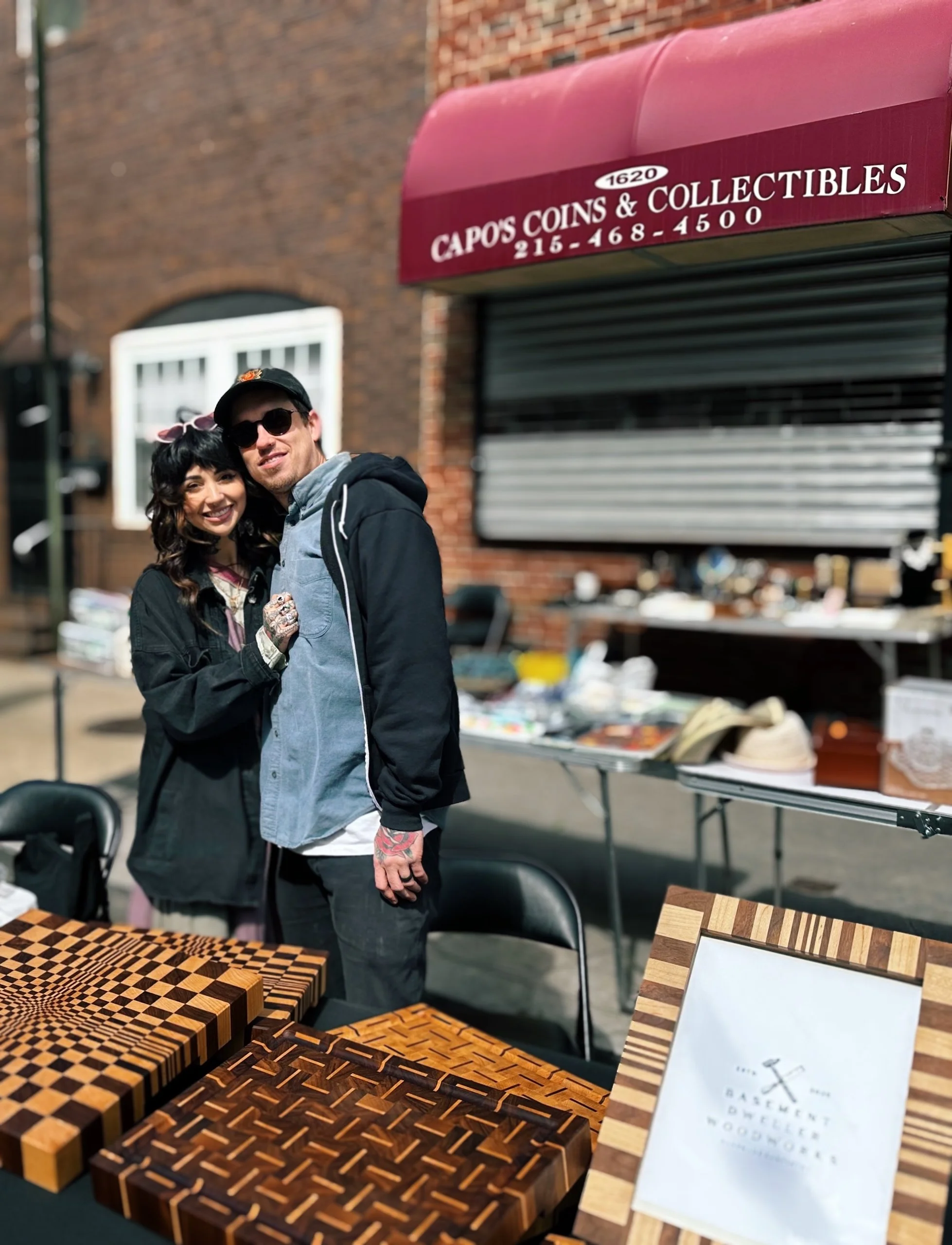 Two people smiling at an outdoor market stall with wooden chessboards displayed on a table and a sign for Capo's Coins & Collectibles in the background.