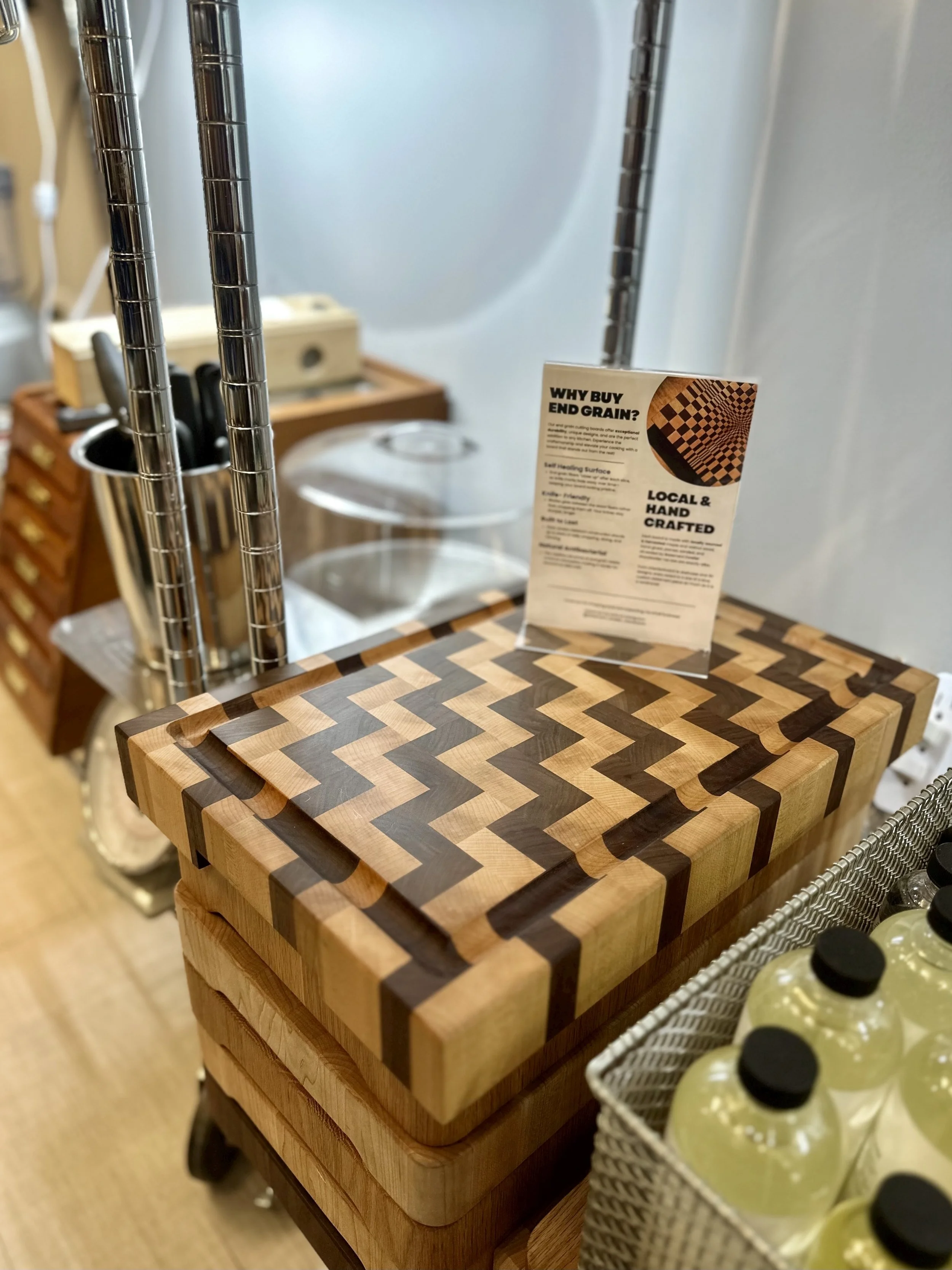 A wooden cutting board with a zigzag patterned inlay, placed on a cart with a metal frame. A small sign about end grain cutting boards stands on top of the cutting board. The background includes knives in a holder, a plastic container, and bottles of hand sanitizer.