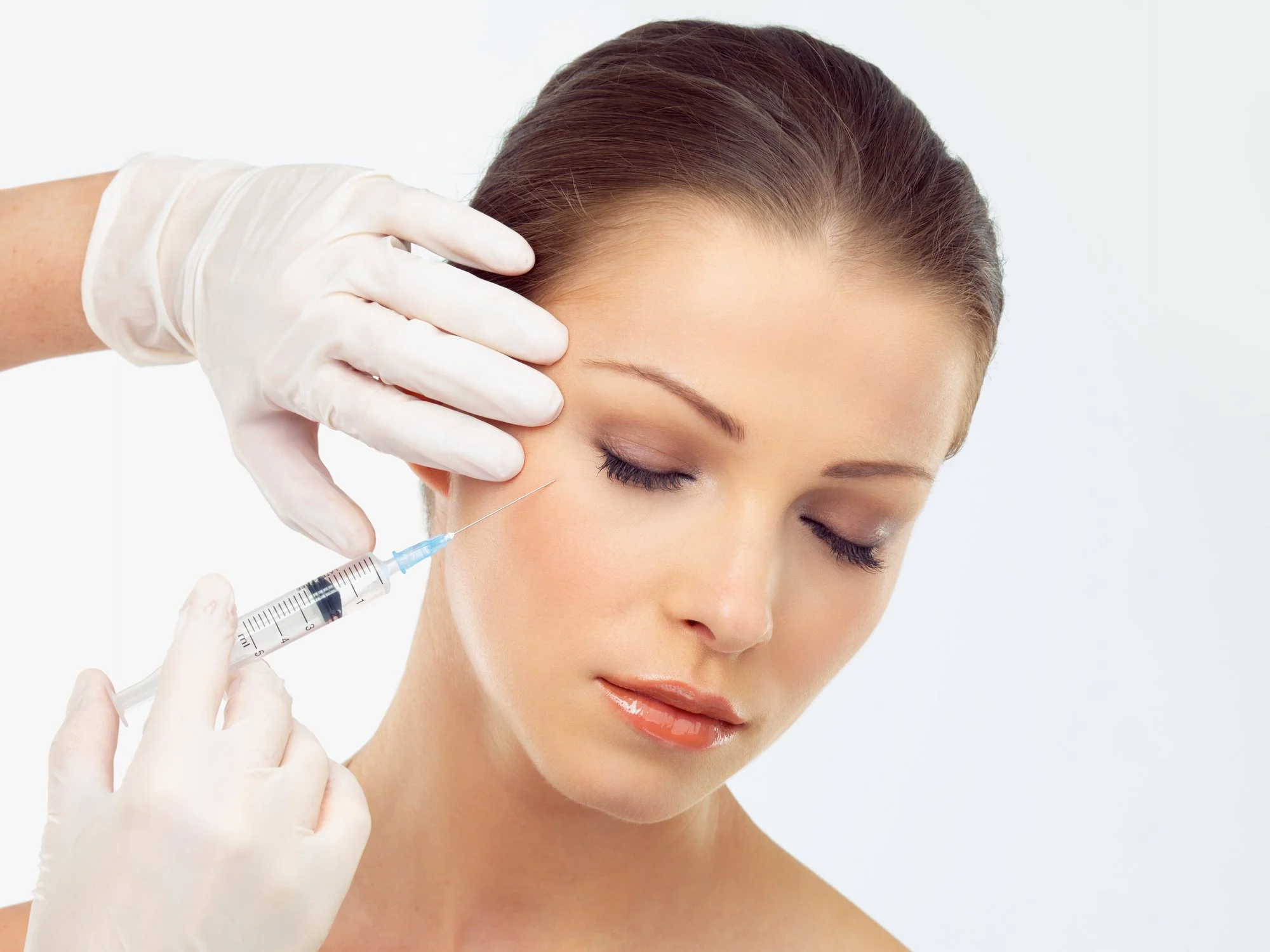 A woman with closed eyes receiving a cosmetic injection in her cheek from a healthcare professional wearing white gloves.