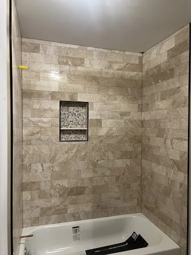 Bathroom shower with beige tiled walls, a built-in recessed shelf with a patterned tile interior, and a white bathtub.