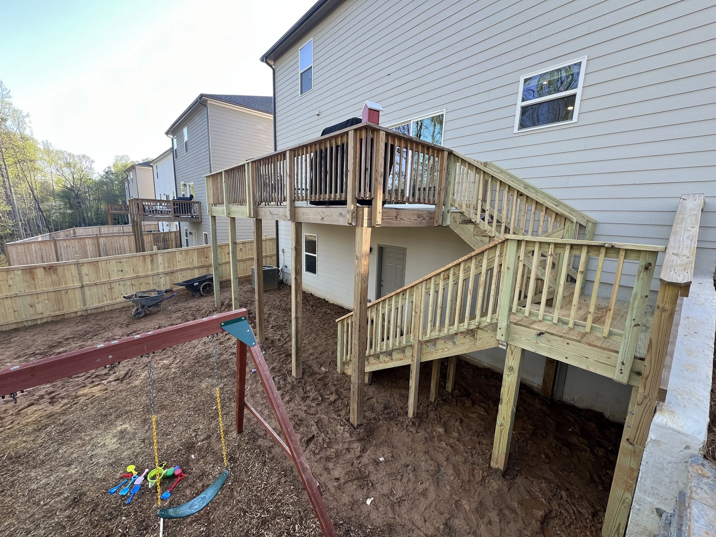 Construction of a multi-level wooden deck with stairs on the back of a beige house, a swing set with toys, and a partially fenced backyard with dirt ground.