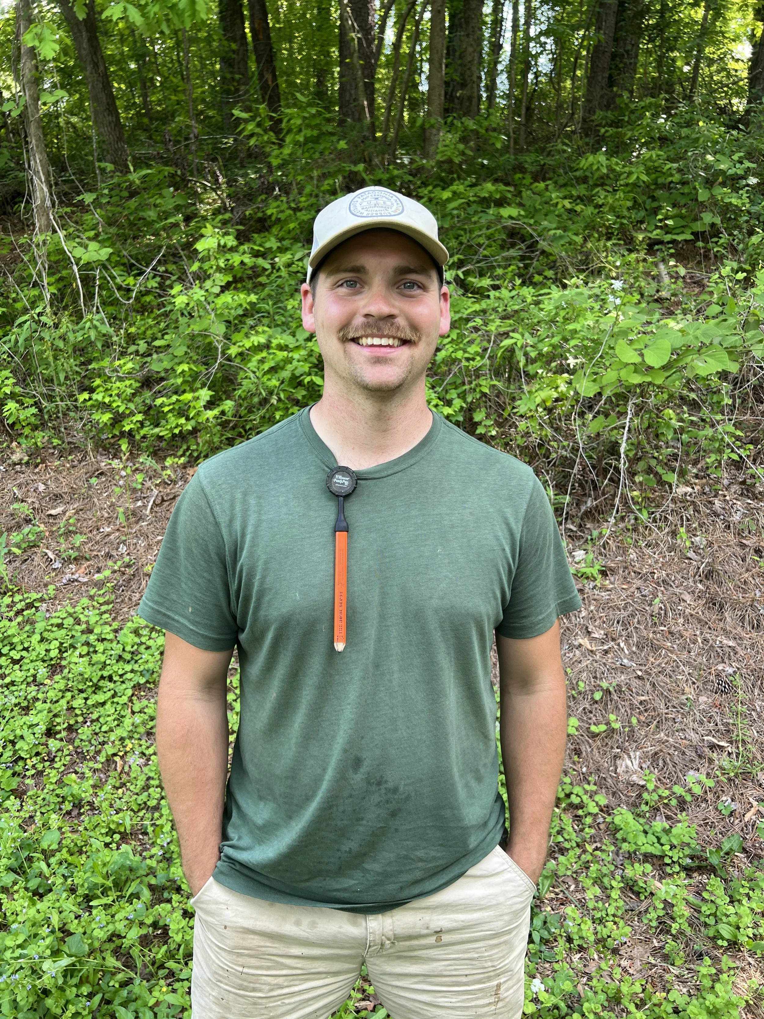 A smiling man with a mustache, wearing a beige cap and green T-shirt, standing outdoors in front of dense green foliage and trees.