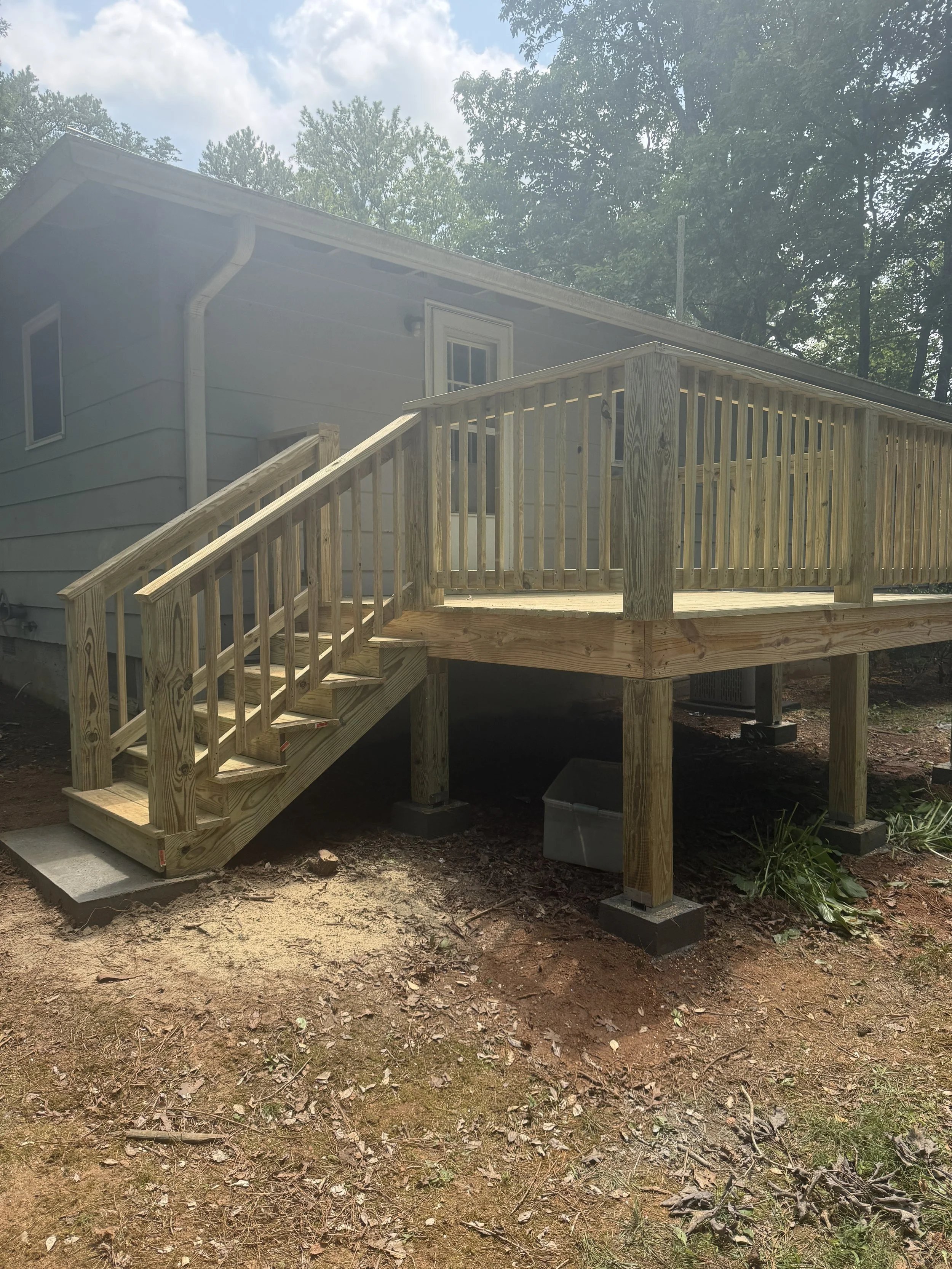 A newly built wooden deck attached to the back of a house, with stairs leading down to dirt ground and surrounded by trees.