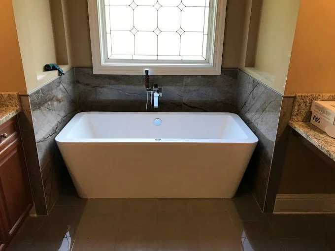 Modern white bathtub positioned beneath a decorative window in a bathroom with marble and tile accents.