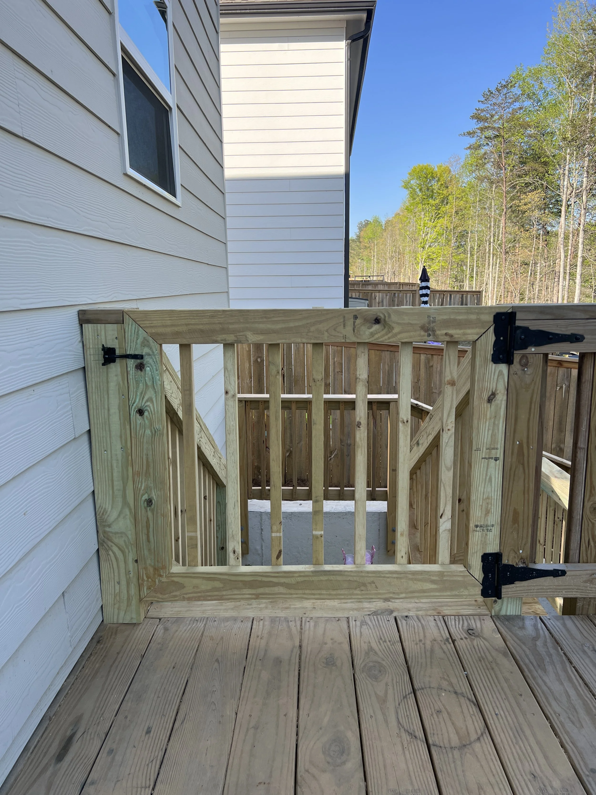 Back porch view showing a newly constructed wooden railing and stairs, with a white house wall and a wooded background.