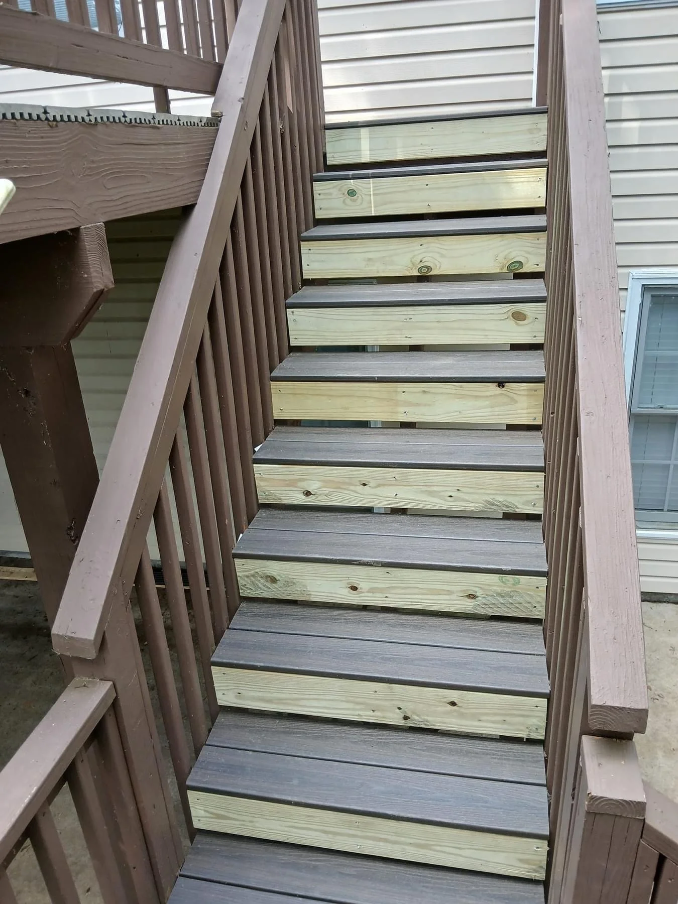 Newly renovated outdoor wooden staircase with contrasting light and dark wood steps and brown railing, attached to a beige house with vinyl siding and a window.