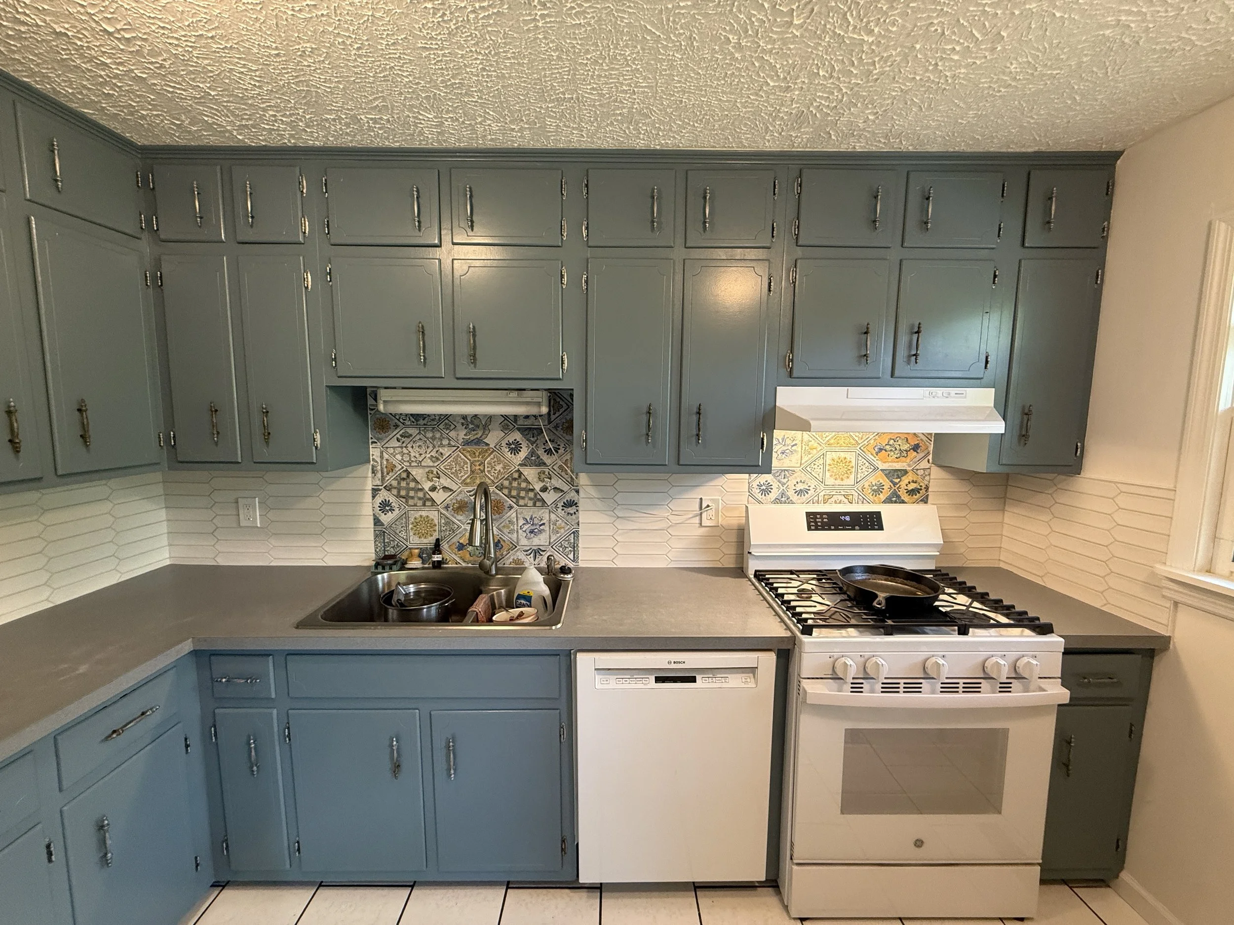 A kitchen with blue lower cabinets and matching upper cabinets, a white stove, white dishwasher, and a gray countertop. There is a colorful tiled backsplash behind the sink and stove, and the floor has hexagon-shaped tiles.