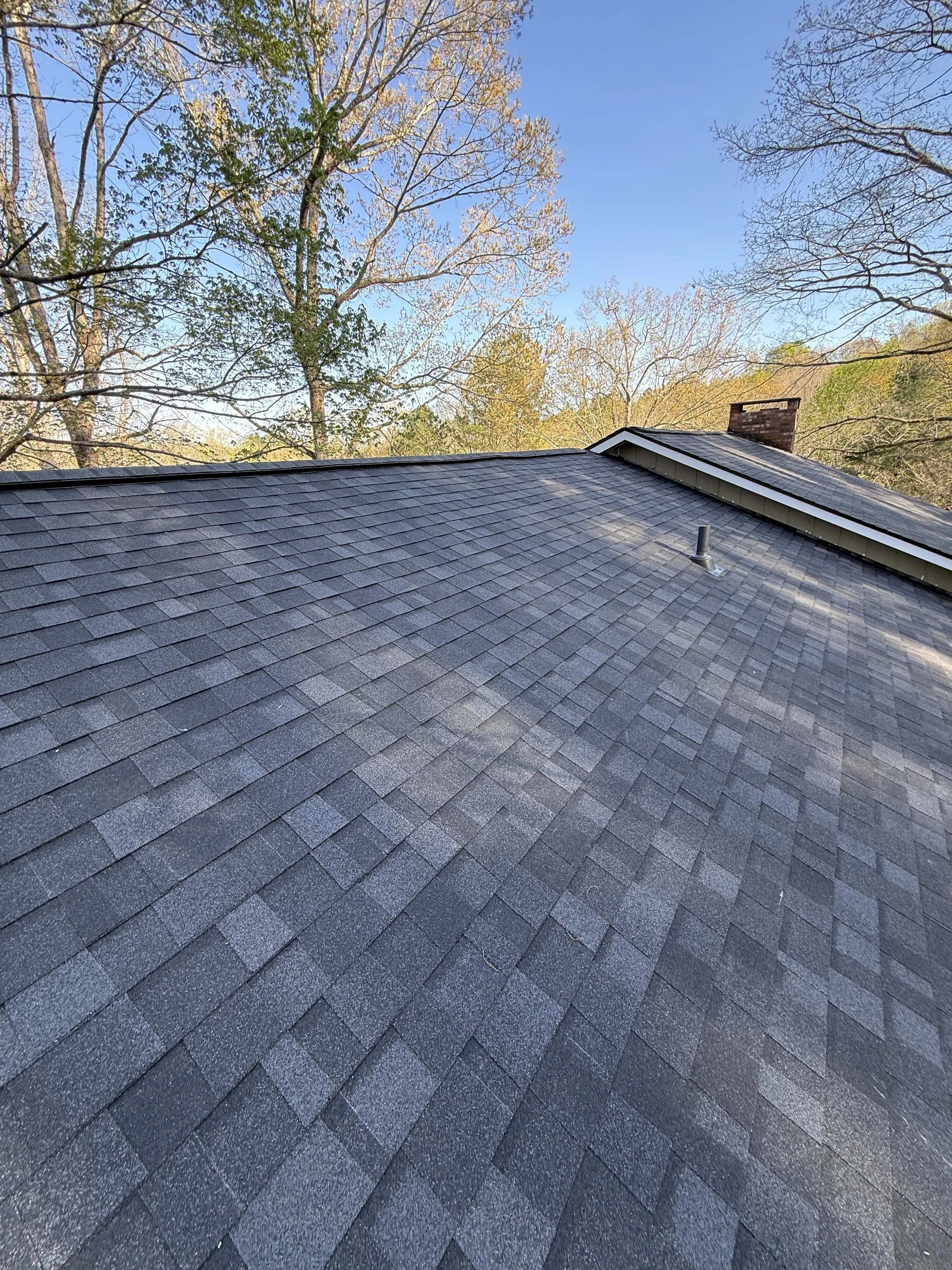 A residential roof with gray shingles, a chimney, and trees in the background against a clear blue sky.