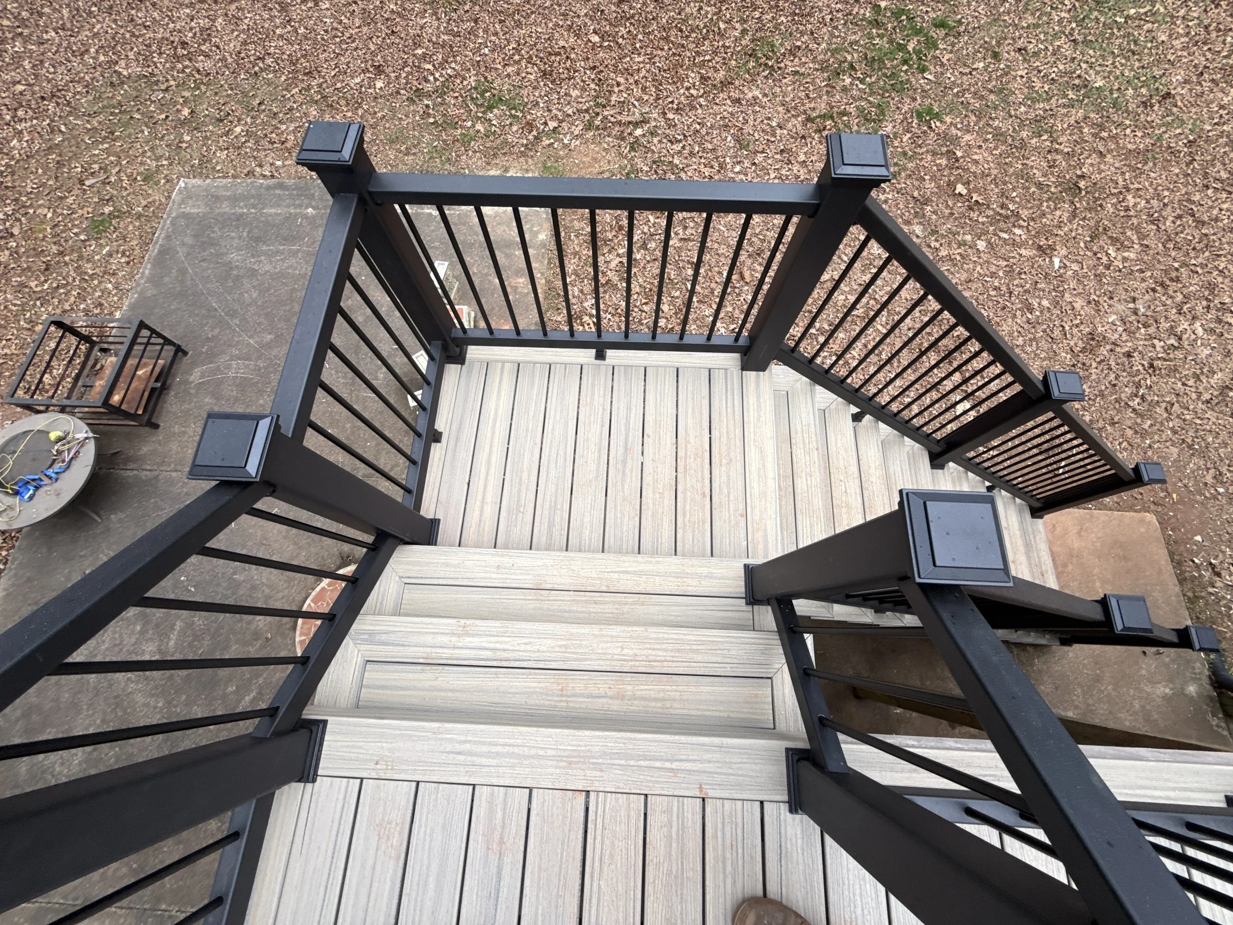 Top-down view of a black metal railing on a wooden deck, with a concrete surface, a small birdcage, and a gravel area with scattered leaves.