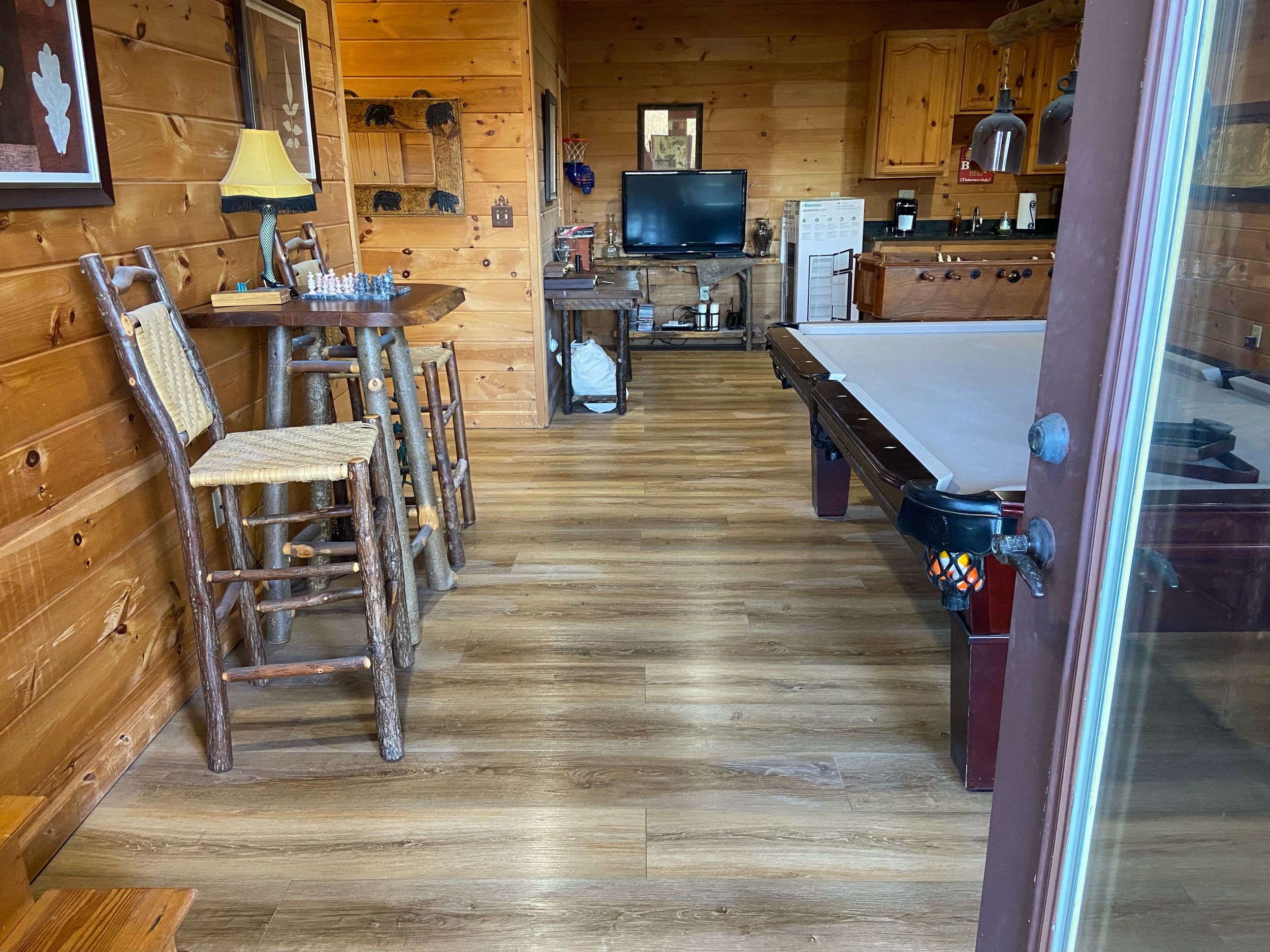 A cozy rustic room with wood-paneled walls and flooring, featuring a high-top table with four log-style chairs, a small TV on a stand, a foosball table, and a kitchenette area with cabinets, a microwave, a coffee maker, and a mini-fridge.