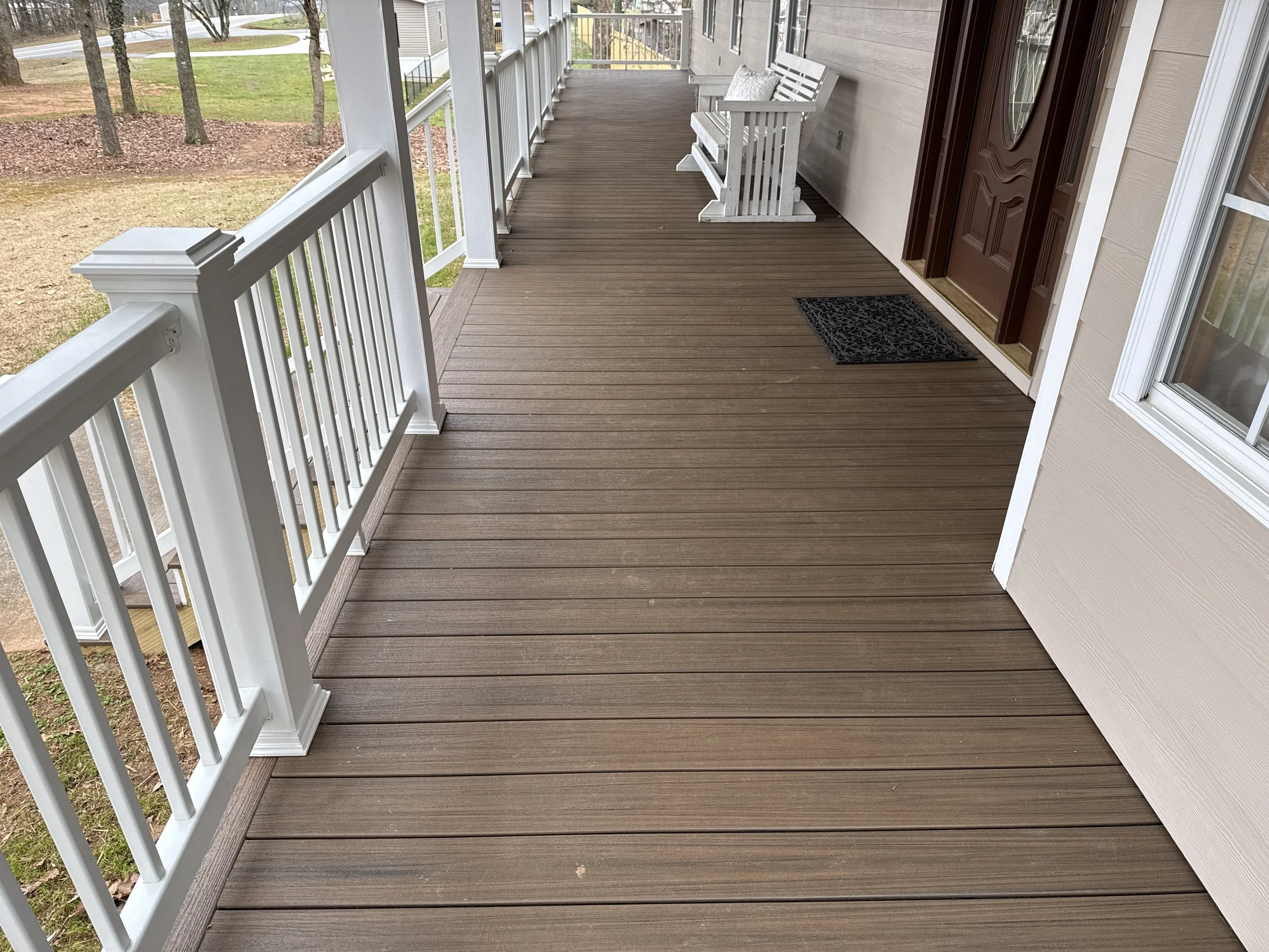 A wooden porch with white railings and a white bench, with a brown front door and a black doormat, overlooking a yard with trees and grass.