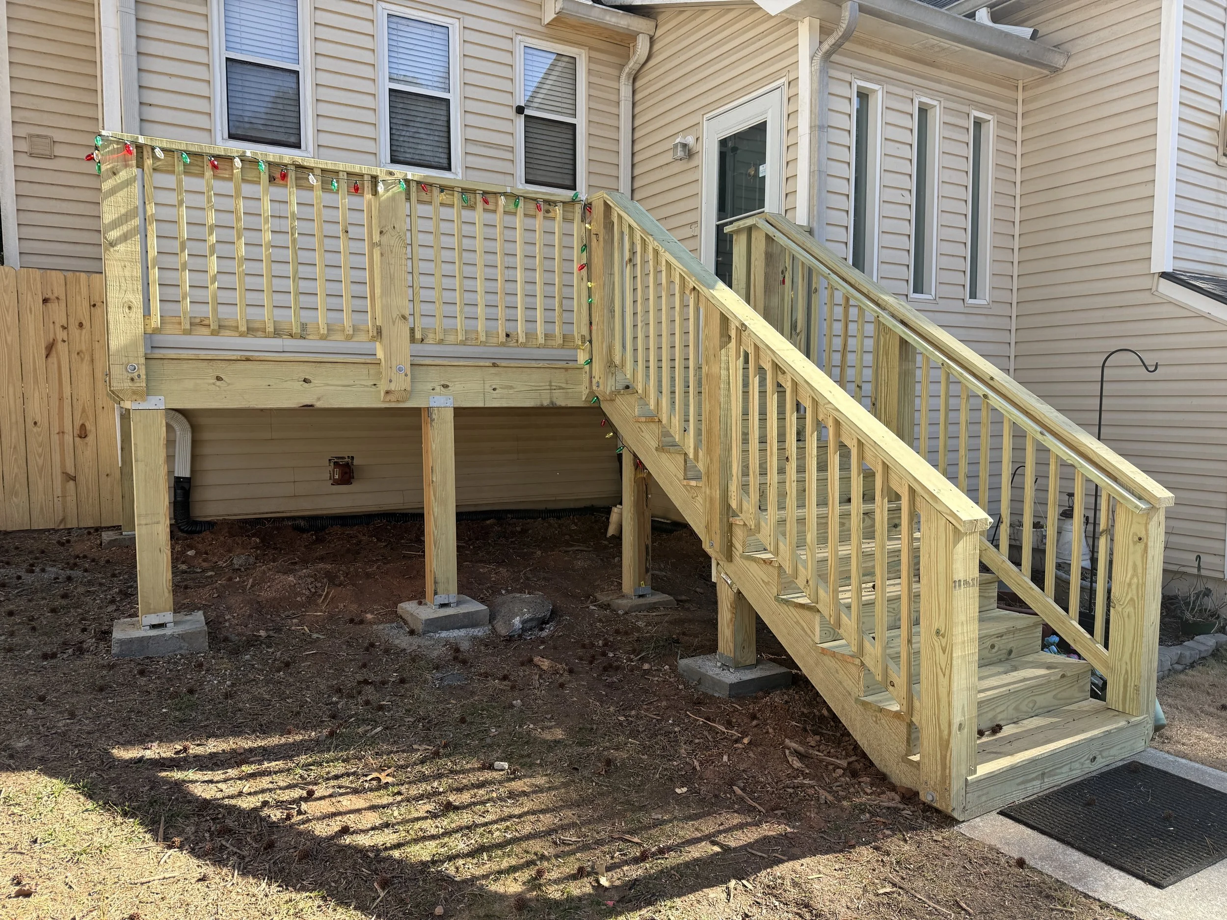 Newly built wooden deck with stairs and railing, decorated with holiday lights, attached to a beige house with multiple windows.