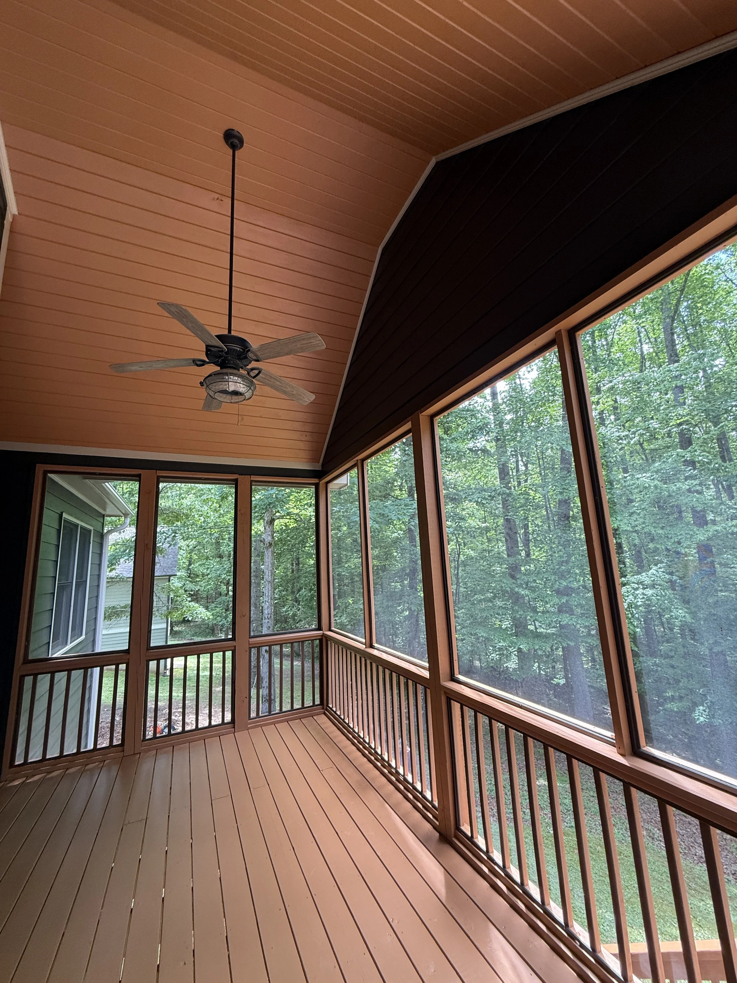 A screened porch with natural wood flooring, a wooden ceiling with a black ceiling fan, large windows with views of green trees outside.