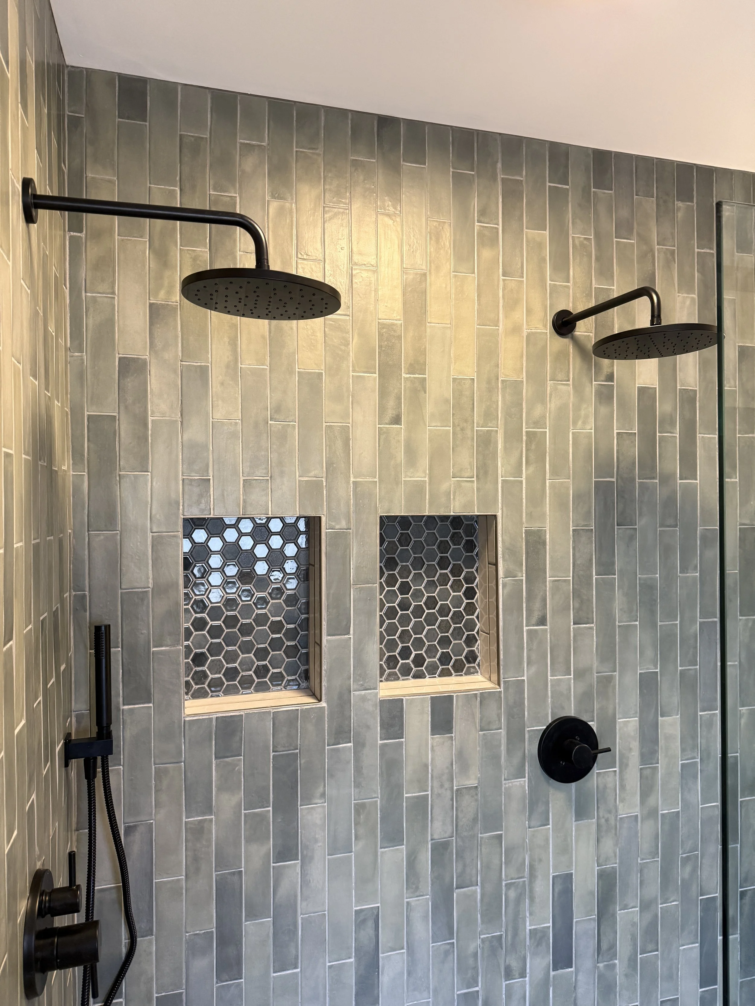 A modern shower with two black rain shower heads mounted on a green-tiled wall, featuring hexagon tile insets.