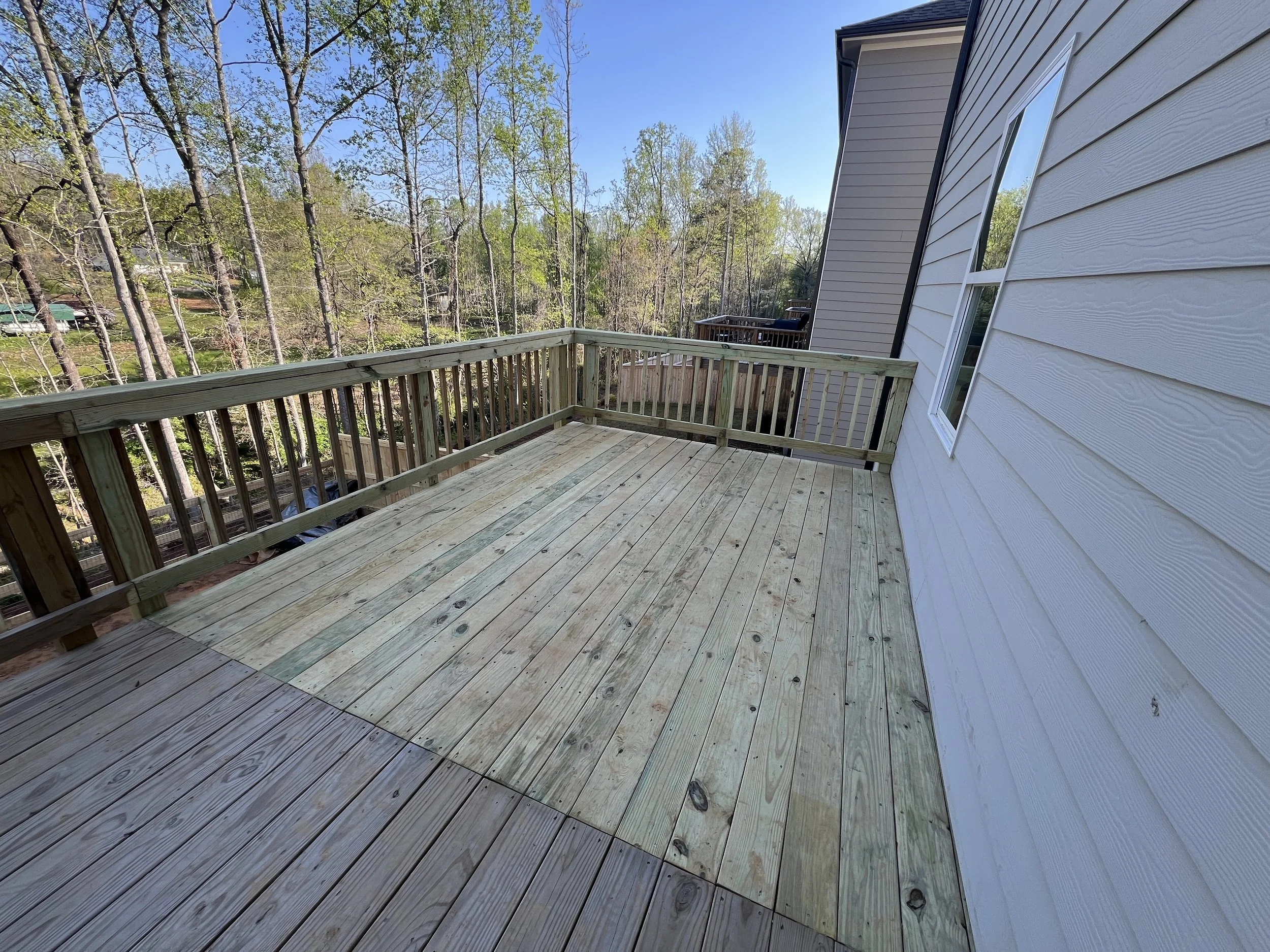 Empty wooden balcony with a railing, attached to a beige house, overlooking a wooded yard with tall trees and neighboring houses.