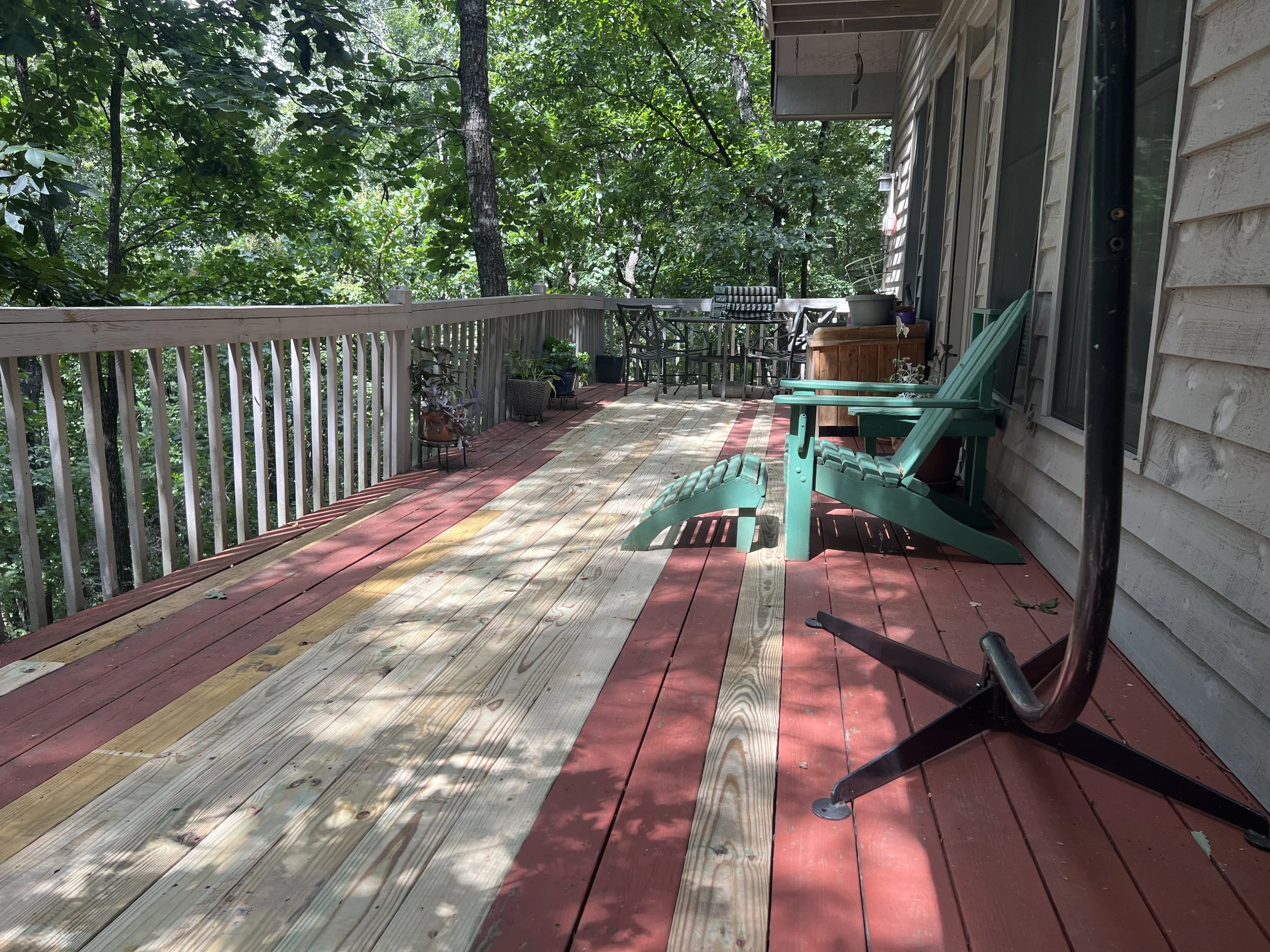 Back porch with red and natural wood decking, green Adirondack chair, and potted plants, overlooking trees.