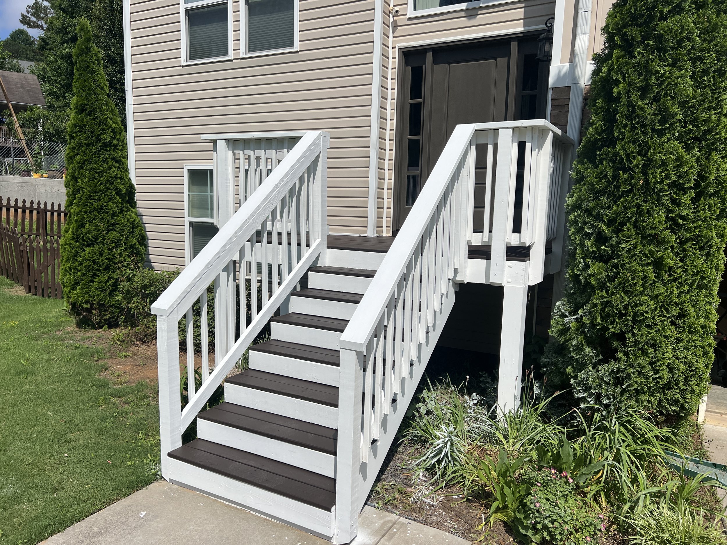 Exterior view of a residential house with a white wooden staircase leading to a side door, surrounded by green bushes and a lawn.