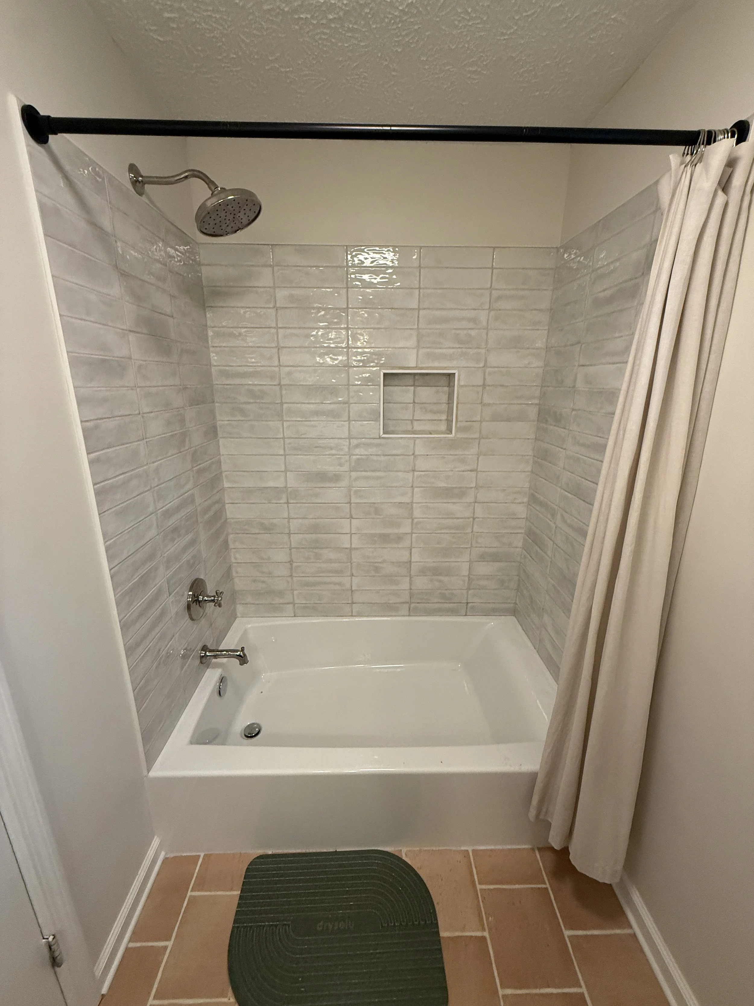 A small shower area with beige tiled walls, a shower head, a built-in shower niche, a soap dish, a curtain to the side, a black striped floor mat, beige floor tiles, and a white tub base.