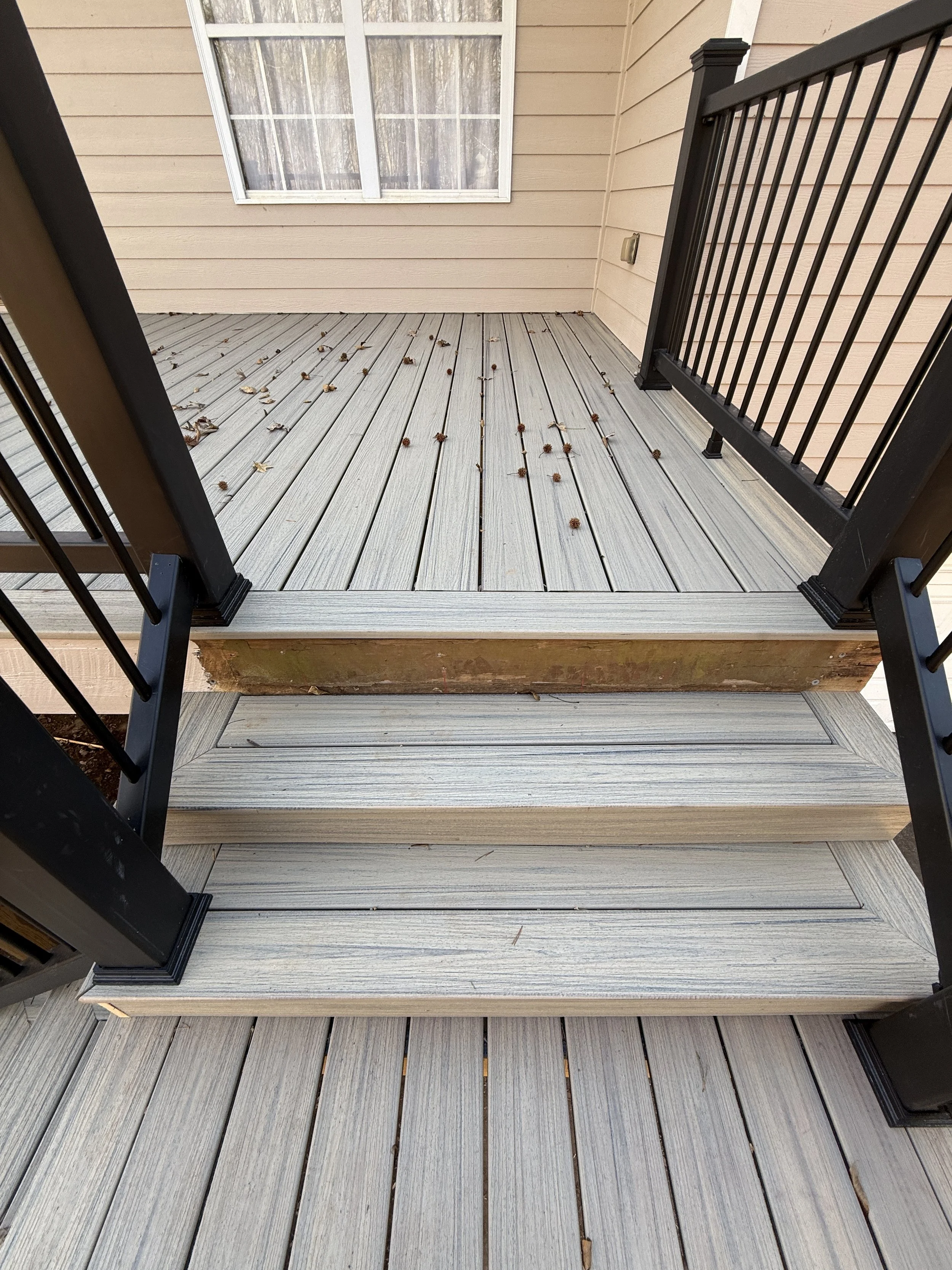 View of a small wooden porch with light grayish composite decking and a black railing, attached to a house with beige horizontal siding. A window with white trim and sheer curtains is seen in the background. Some fallen leaves and pinecones are scattered on the porch.