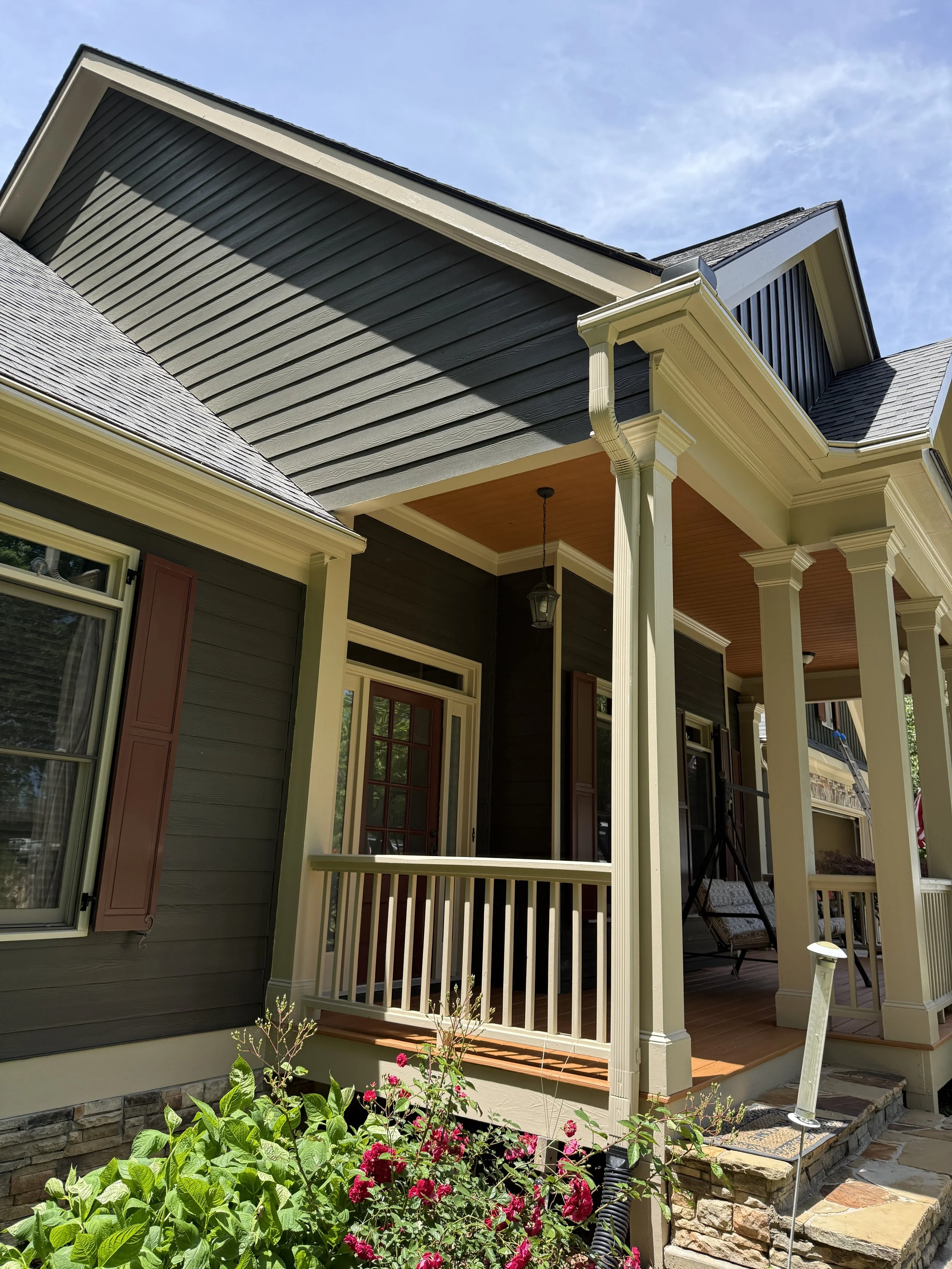 View of a house porch with decorative columns, dark gray siding, a small balcony with a white railing, and vibrant pink and green garden plants in the foreground, under a blue sky.