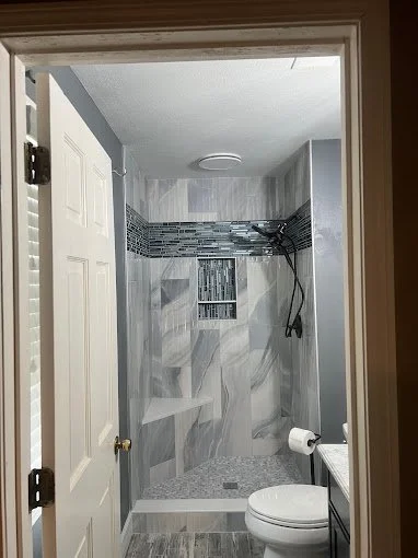 View through bathroom doorway showing a tiled walk-in shower with built-in shelf, a handheld shower head, and a toilet with a roll of toilet paper on a nearby holder.