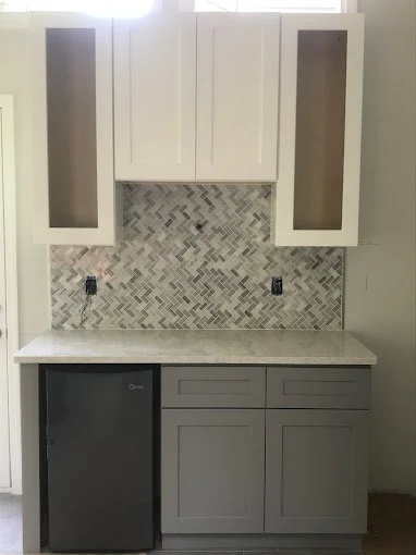 Kitchen upper cabinets and lower cabinets with a small mini fridge, backsplash with gray herringbone tiles, and a marble countertop