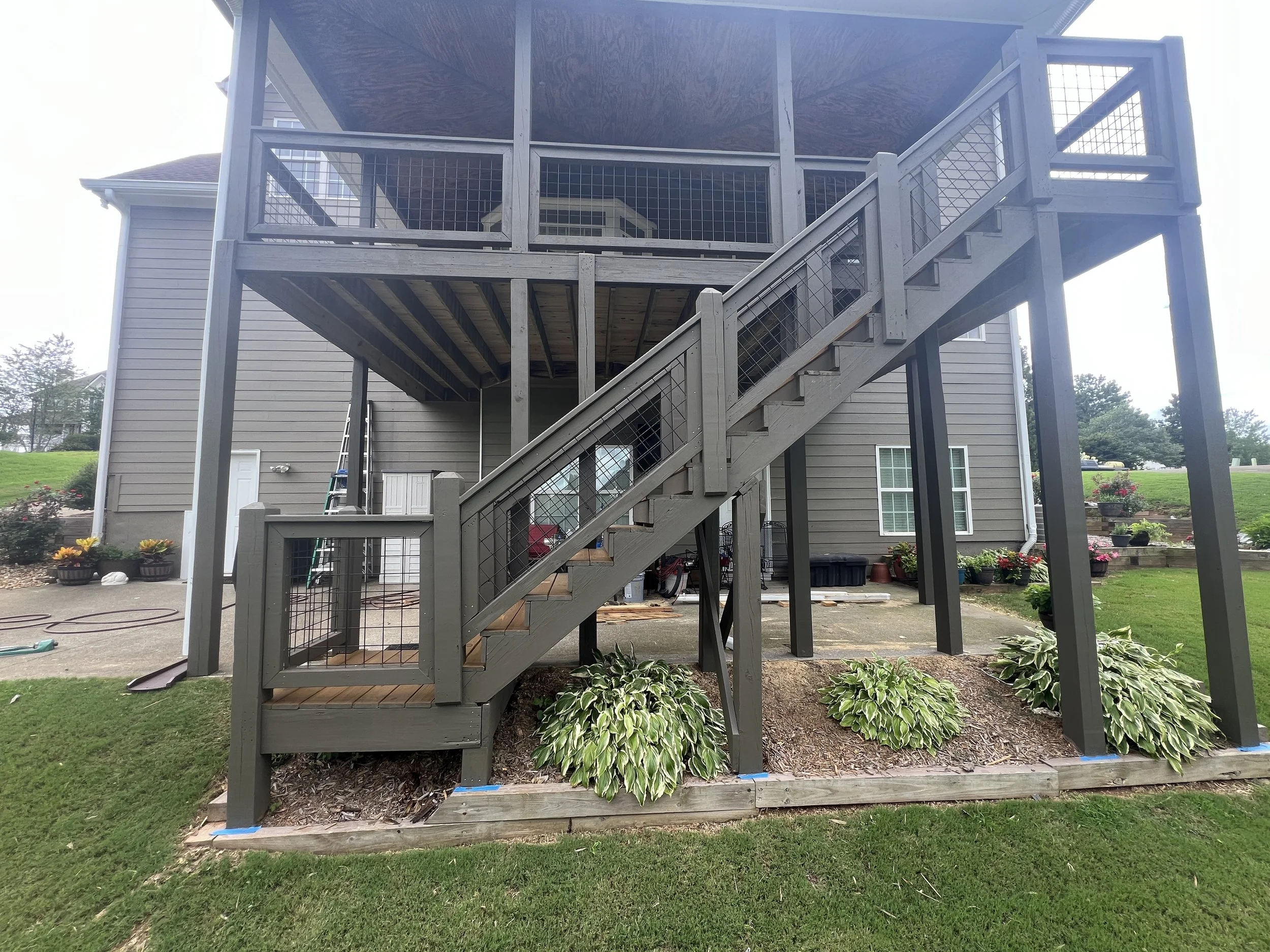 Backyard with a two-story wooden deck attached to a house, accessible by a staircase with a handrail, surrounded by plants, flowers, and a grassy lawn.