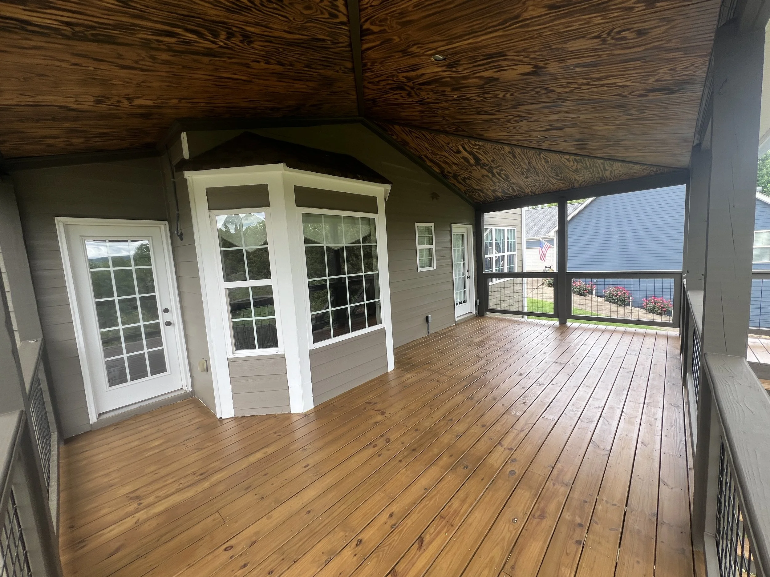 Large covered porch with wooden flooring, black railings, exterior walls, and windows of a house, overlooking a neighborhood yard with flowers.