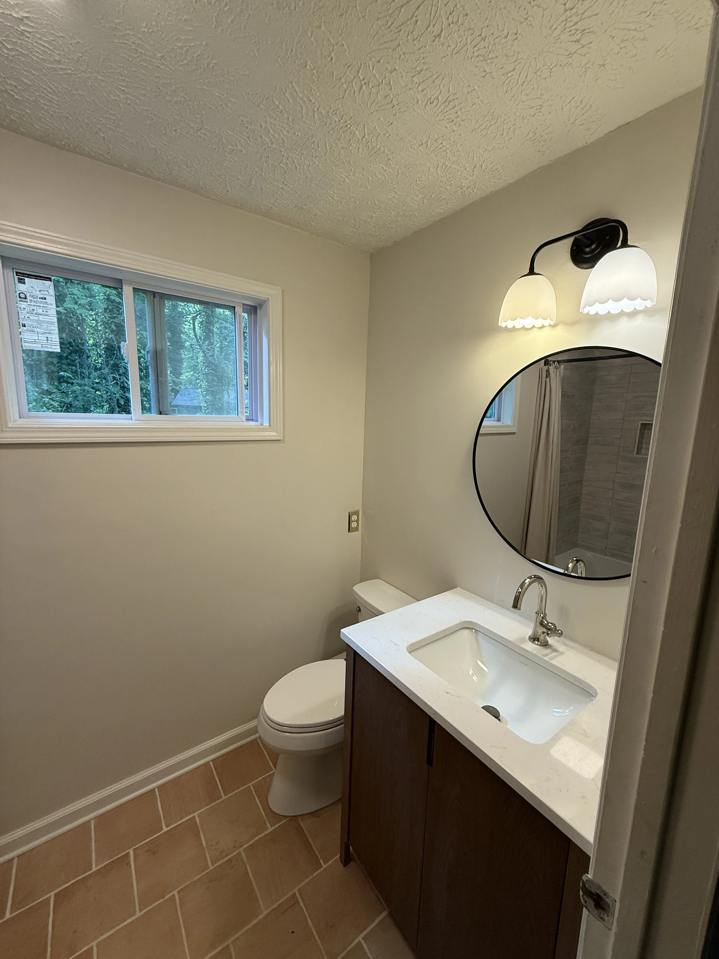 A small bathroom with a window showing surrounding trees, a wall-mounted light fixture with two lamps, a round mirror, a white sink with a dark wood cabinet, a toilet, and beige tiled floors.