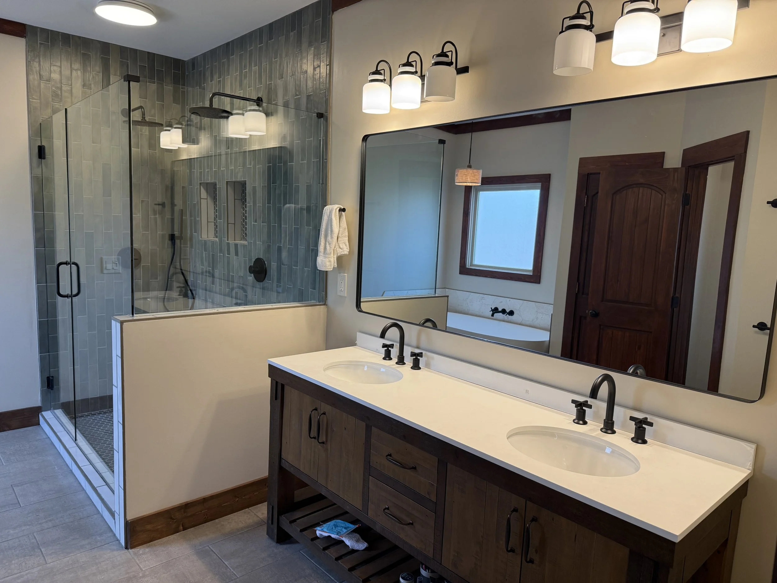 Modern bathroom with double vanity, large mirror, and walk-in shower with glass enclosure. The vanity has two sinks with black fixtures, and a wooden cabinet below. The lighting above the mirror features multiple white shades. There is a window and wooden door in the background.